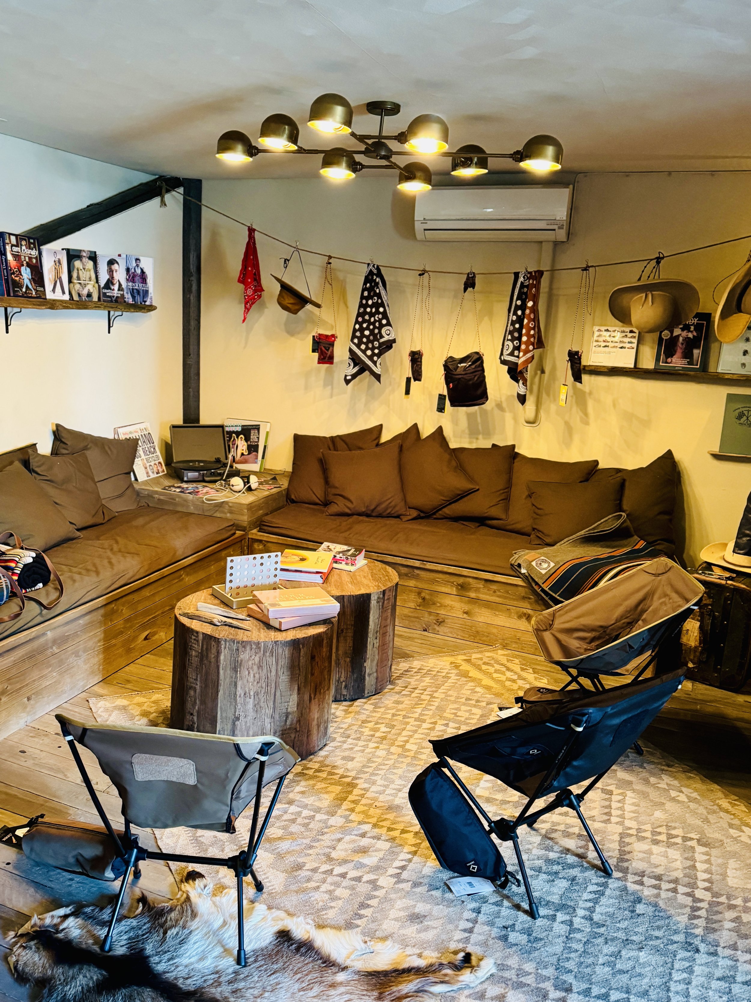Cozy rustic living room with a corner sofa, wooden coffee tables, folding chairs, and decorative items hanging on a line. Shelves display books and hats. The floor features a patterned rug and a fur rug. A modern light fixture hangs from the ceiling.