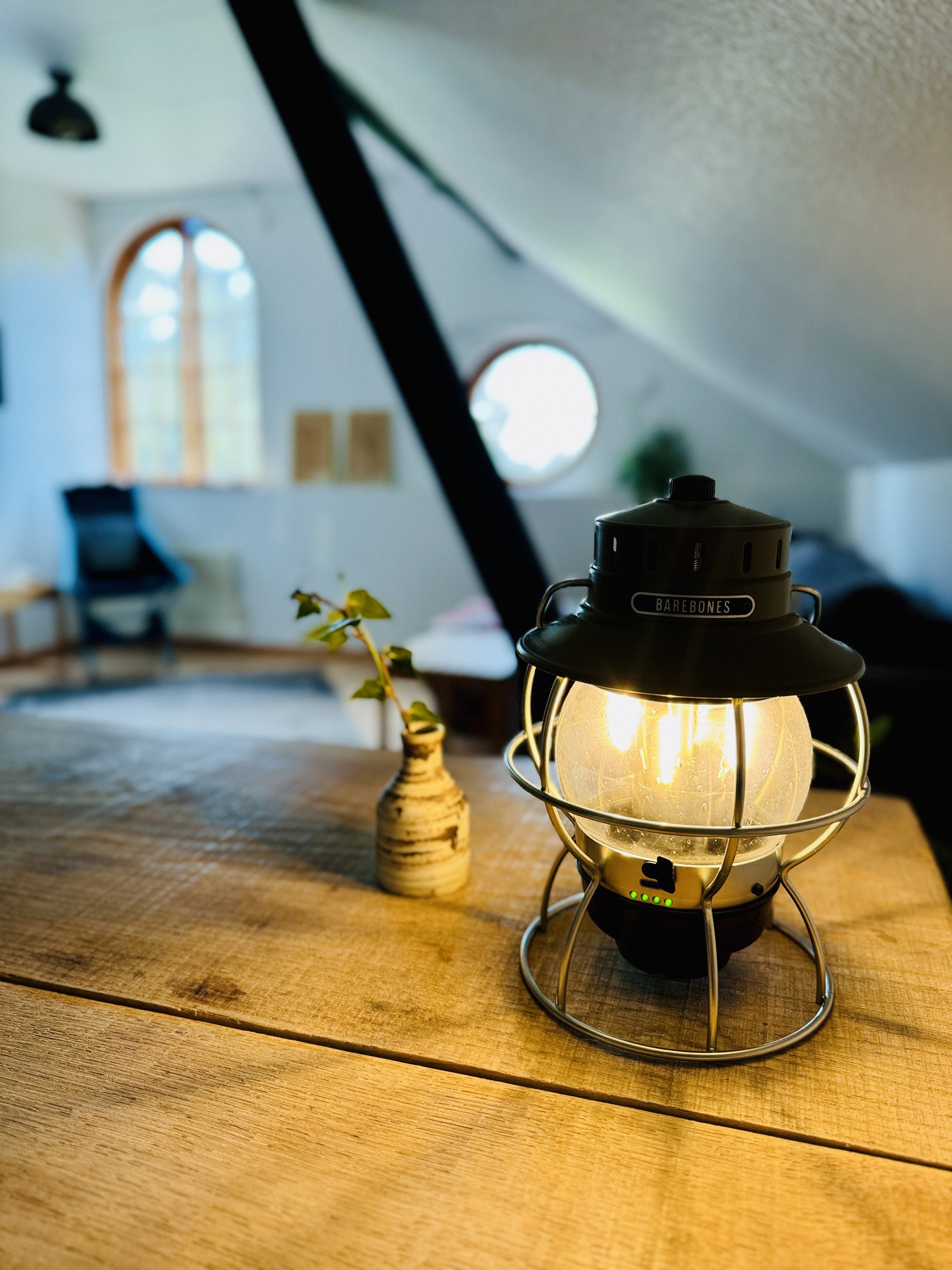 A vintage-style Barebones lantern on a wooden table with a small vase containing a plant in a cozy room with arched windows.