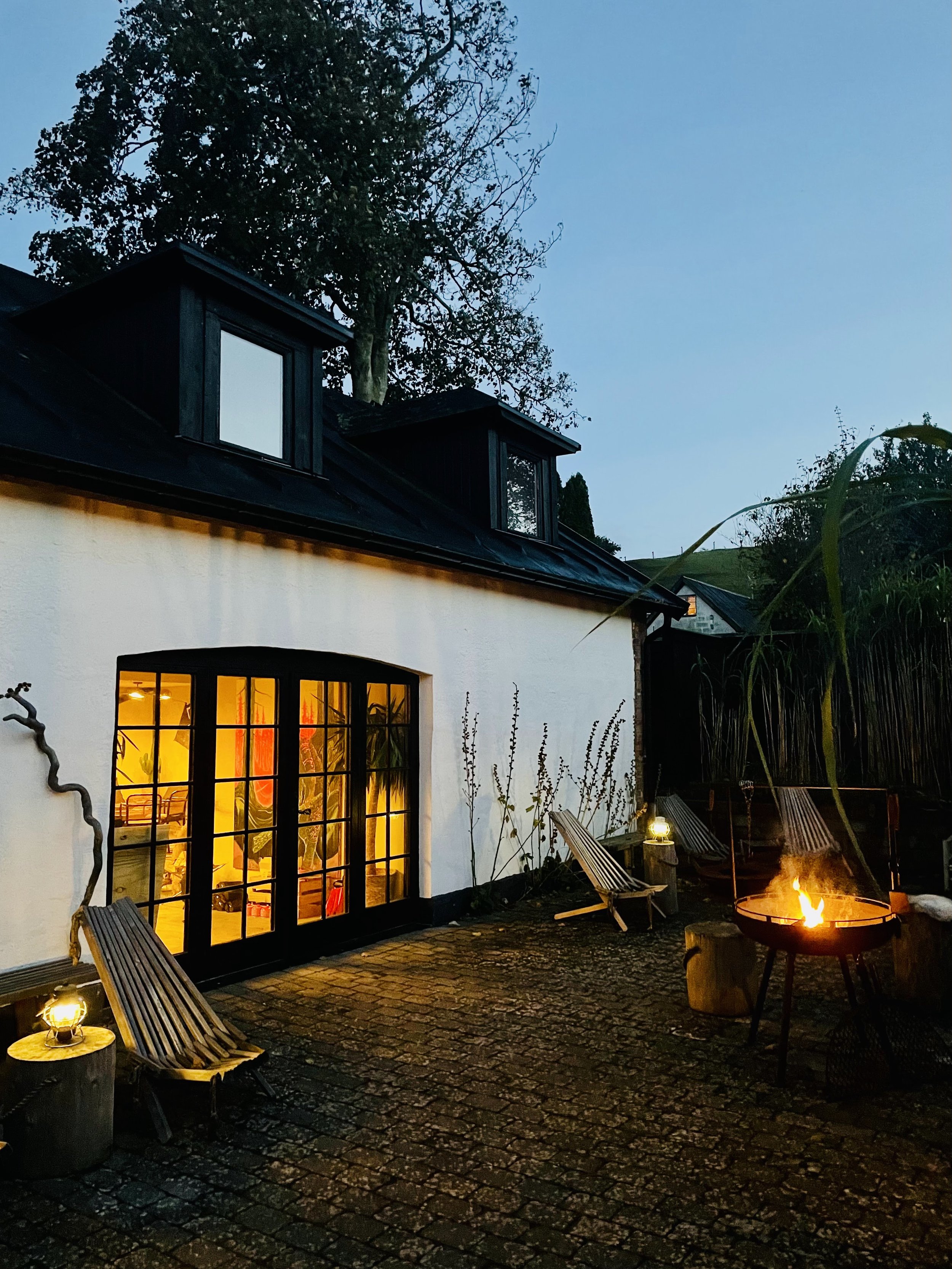 Cozy outdoor scene with a fire pit, chairs, lanterns, and a charming house with lit windows at dusk.
