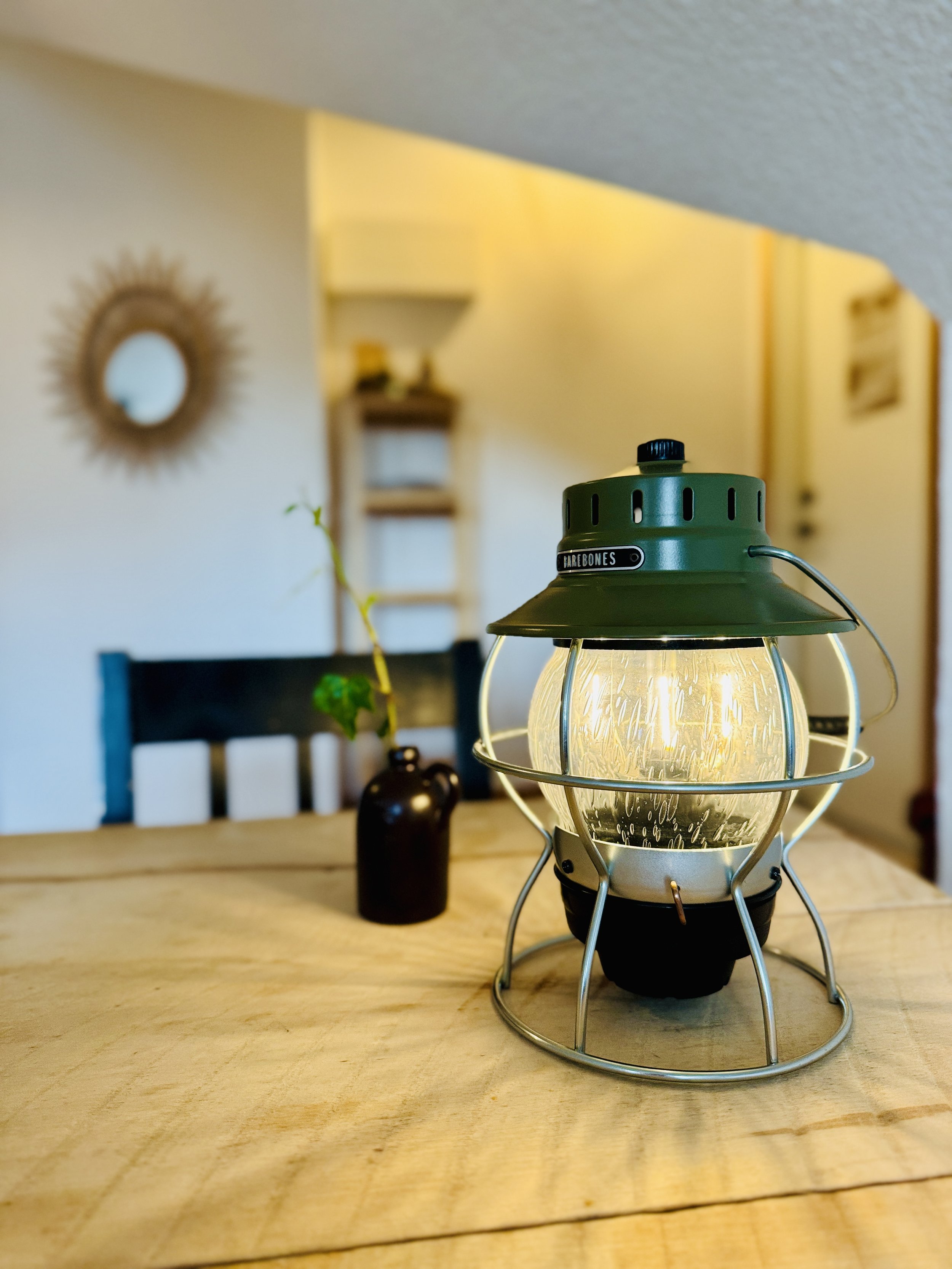 A vintage-style Barebones lantern with a green top sits on a wooden table. In the background, a small dark vase with a plant stem and a decorative mirror are visible. The setting has a cozy indoor atmosphere.