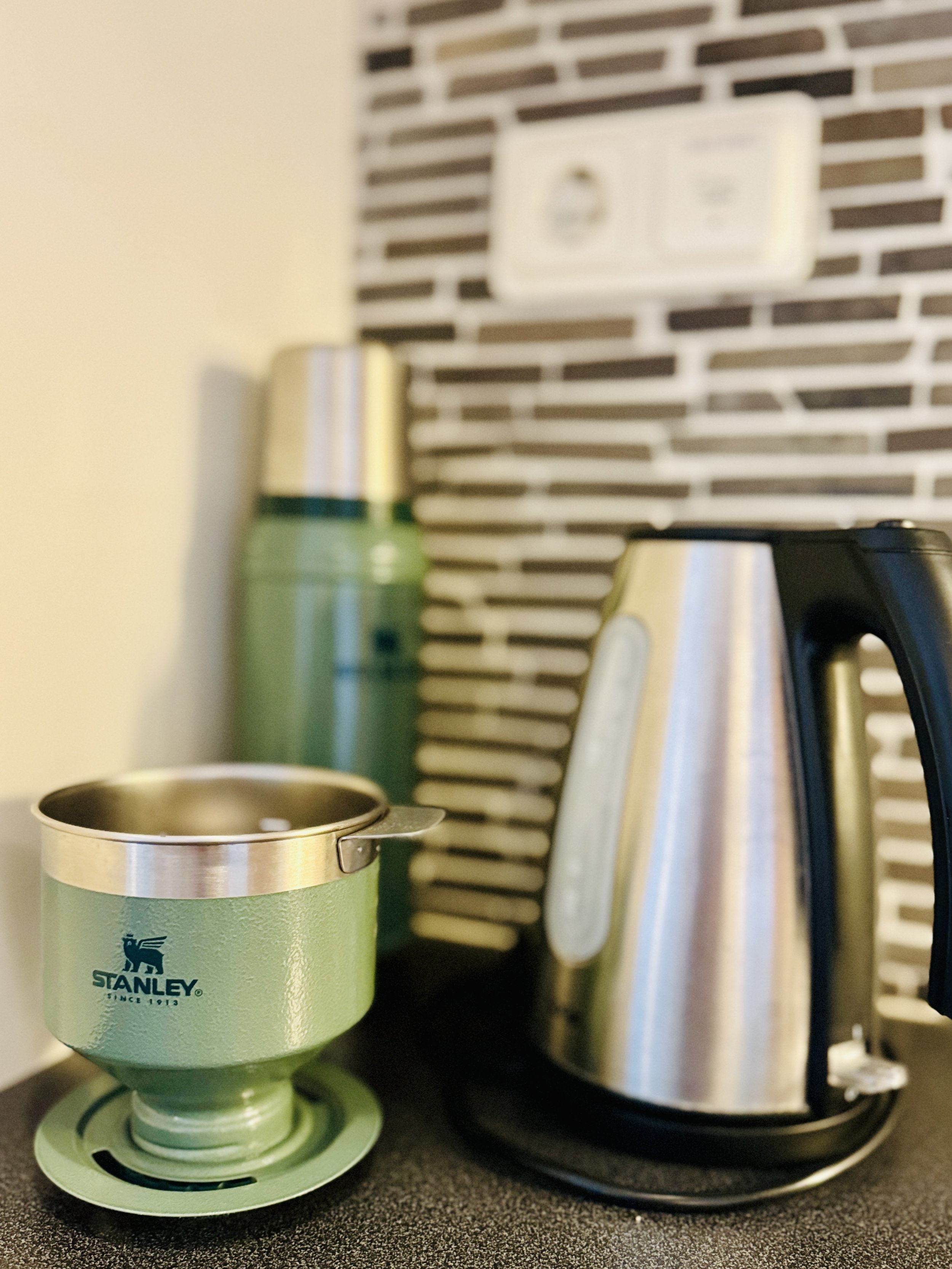 Green Stanley camping coffee percolator, green Stanley thermos, and stainless steel electric kettle on kitchen counter with tile backsplash.