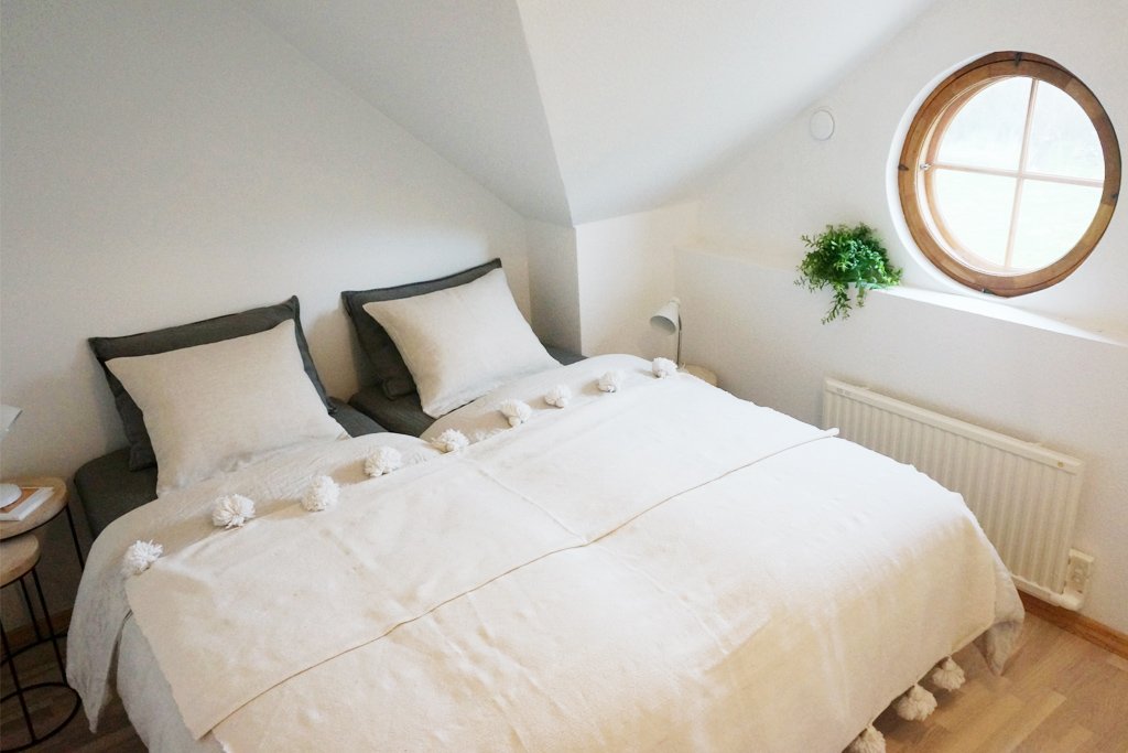 Cozy bedroom interior with a double bed, white bedding, and decorative pillows. The room features a round window, a small green plant on the windowsill, and a modern bedside lamp on a wooden side table. A radiator is visible beneath the window.