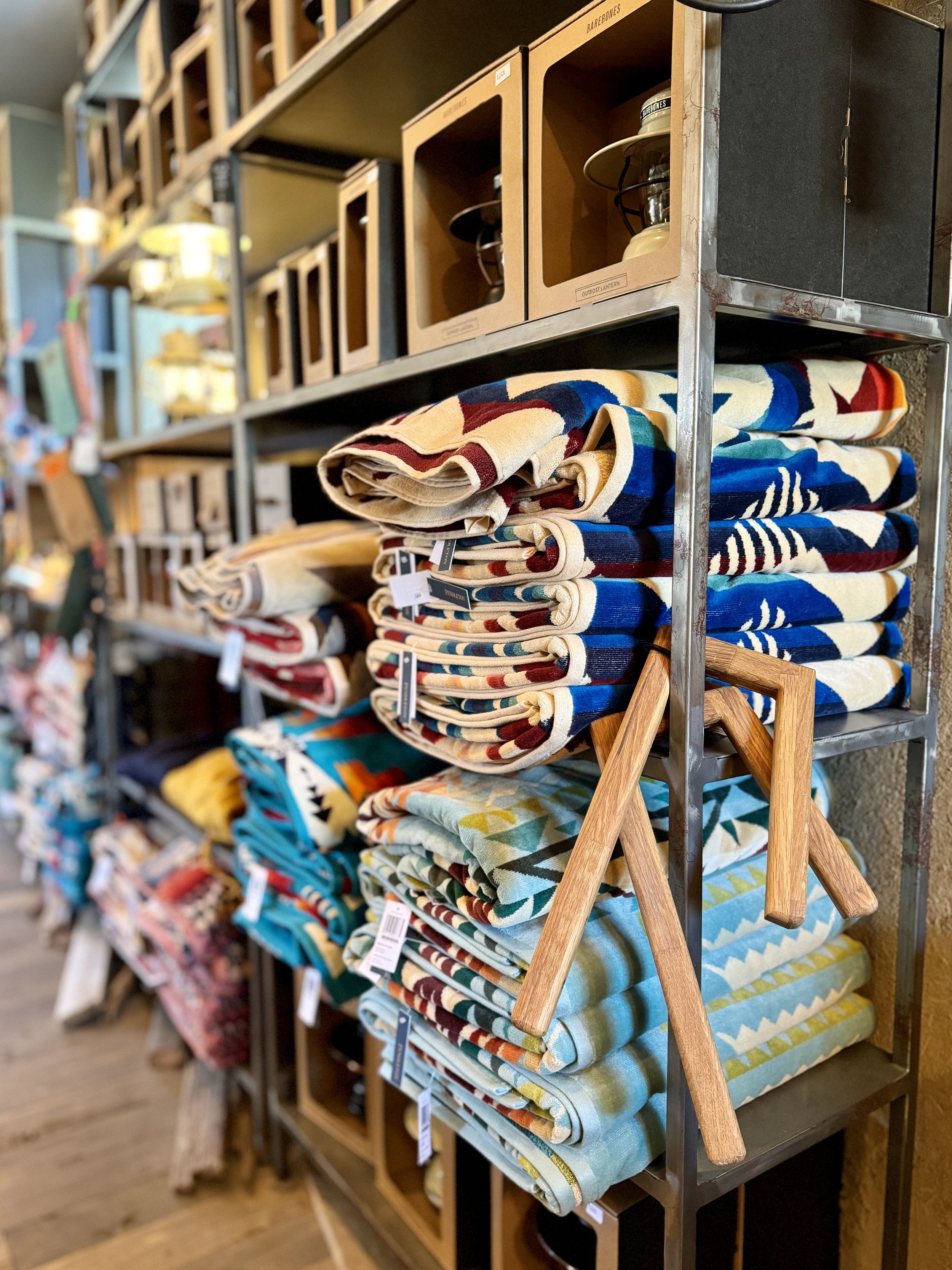 A shelf displaying folded colorful blankets with geometric patterns and wooden hanger handles. Boxes containing lamps are visible on the top shelf.
