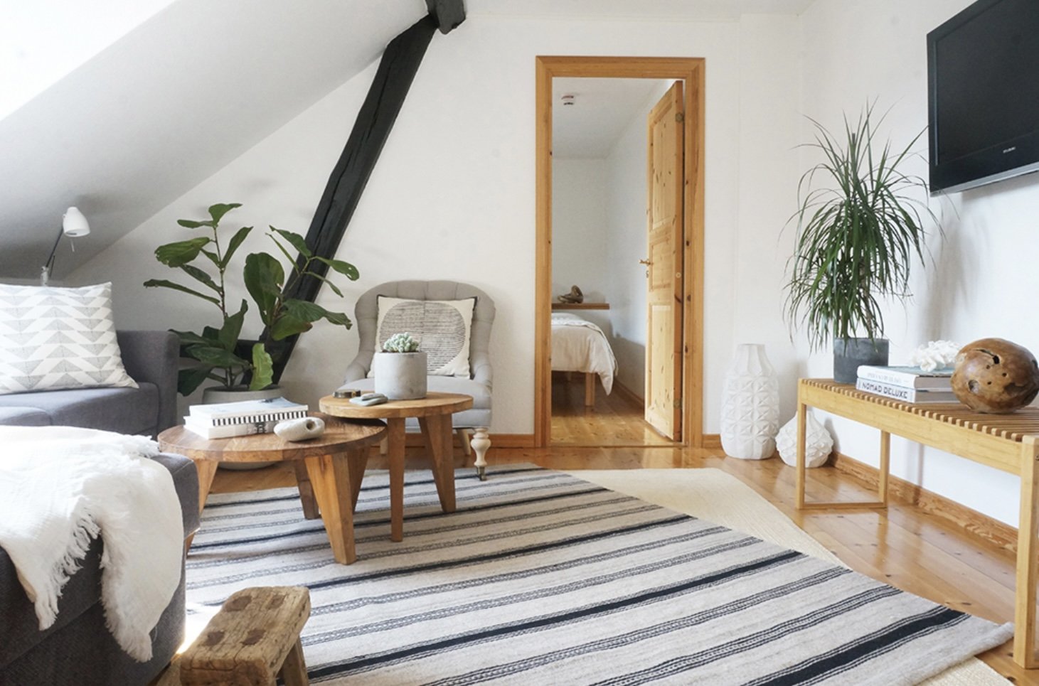 Cozy living room with modern decor, featuring a gray sofa, a striped rug, wooden coffee tables, a potted plant, and wall-mounted TV. A doorway opens to a bedroom with a visible bed.