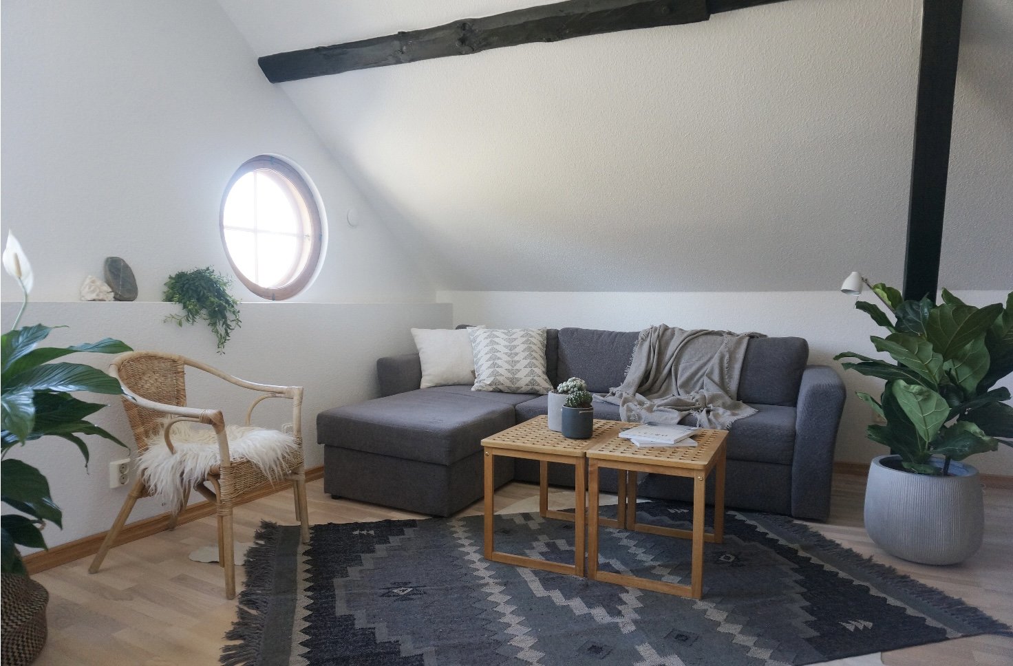 Cozy loft living room with gray sectional sofa, wooden round window, cactus plant on coffee table, wicker chair with white cushion, and decorative plants. Features natural light and wooden beams.