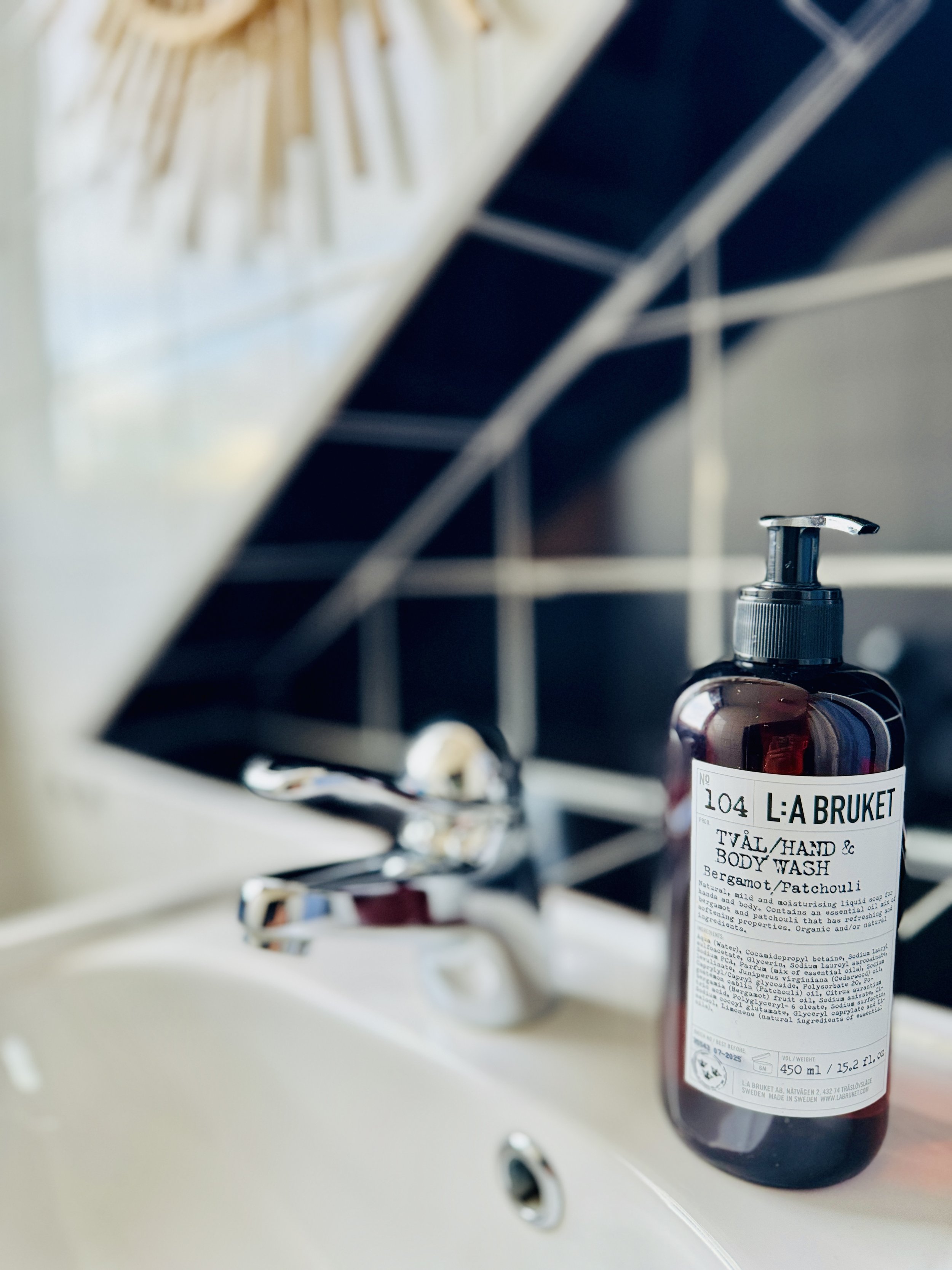 L:A Bruket hand and body wash bottle on a bathroom sink with silver faucet, black and white tile background.