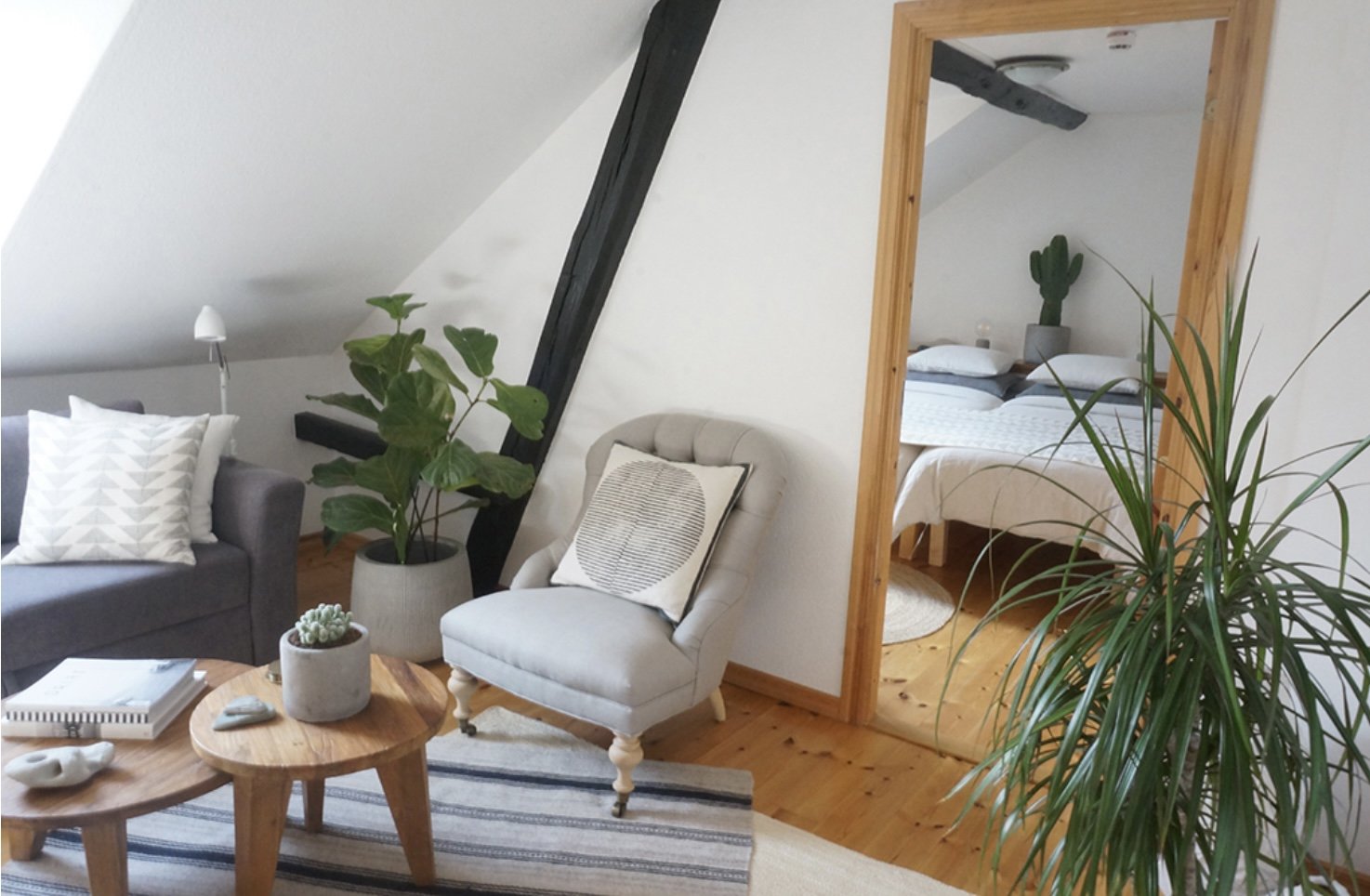 Cozy living room with gray armchair, potted plants, wooden coffee tables, and a gray couch with pillows, wooden flooring, and a mirror reflecting a bed with a cactus on the nightstand.
