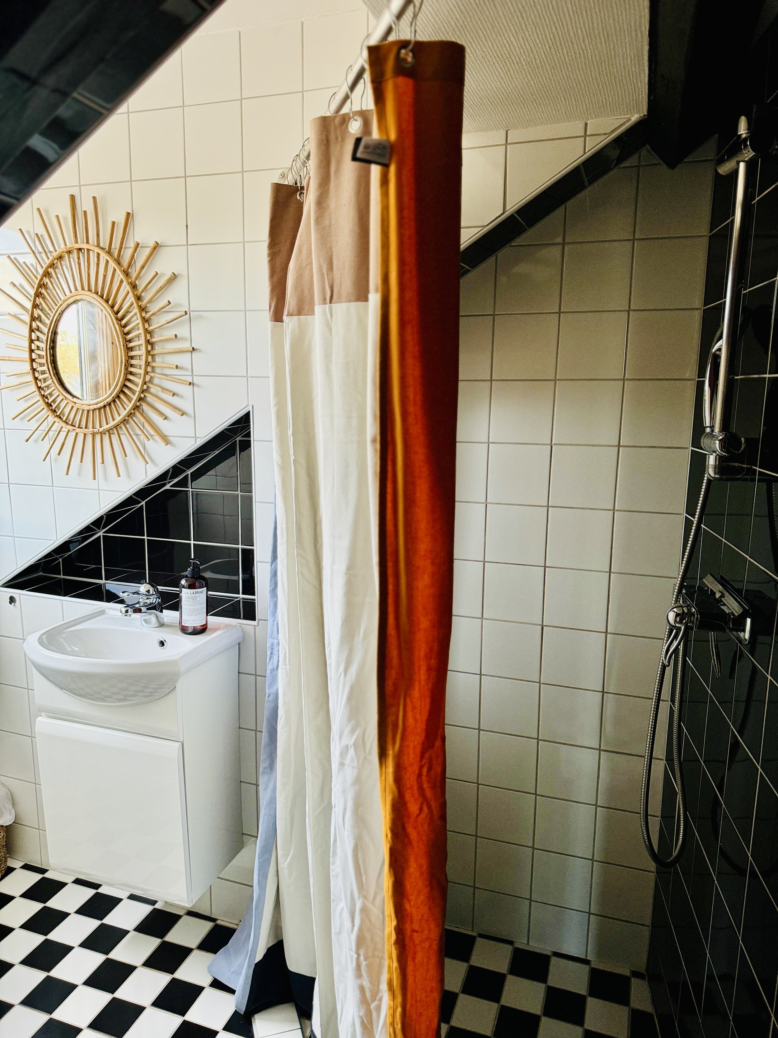 Bathroom with geometric design featuring a black and white checkered floor, white tiled walls, a shower with a multicolored curtain, and a white sink with a round mirror surrounded by a sunburst frame.