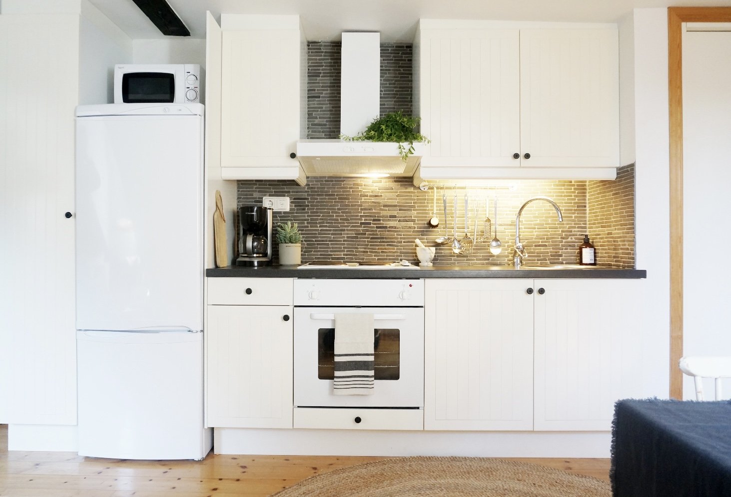 Modern kitchen interior with white cabinets, a refrigerator, oven, stovetop, microwave, and range hood. The countertop features a coffee maker, cutting board, and potted plant. Textured backsplash with utensils hanging. Wooden floor and round rug.