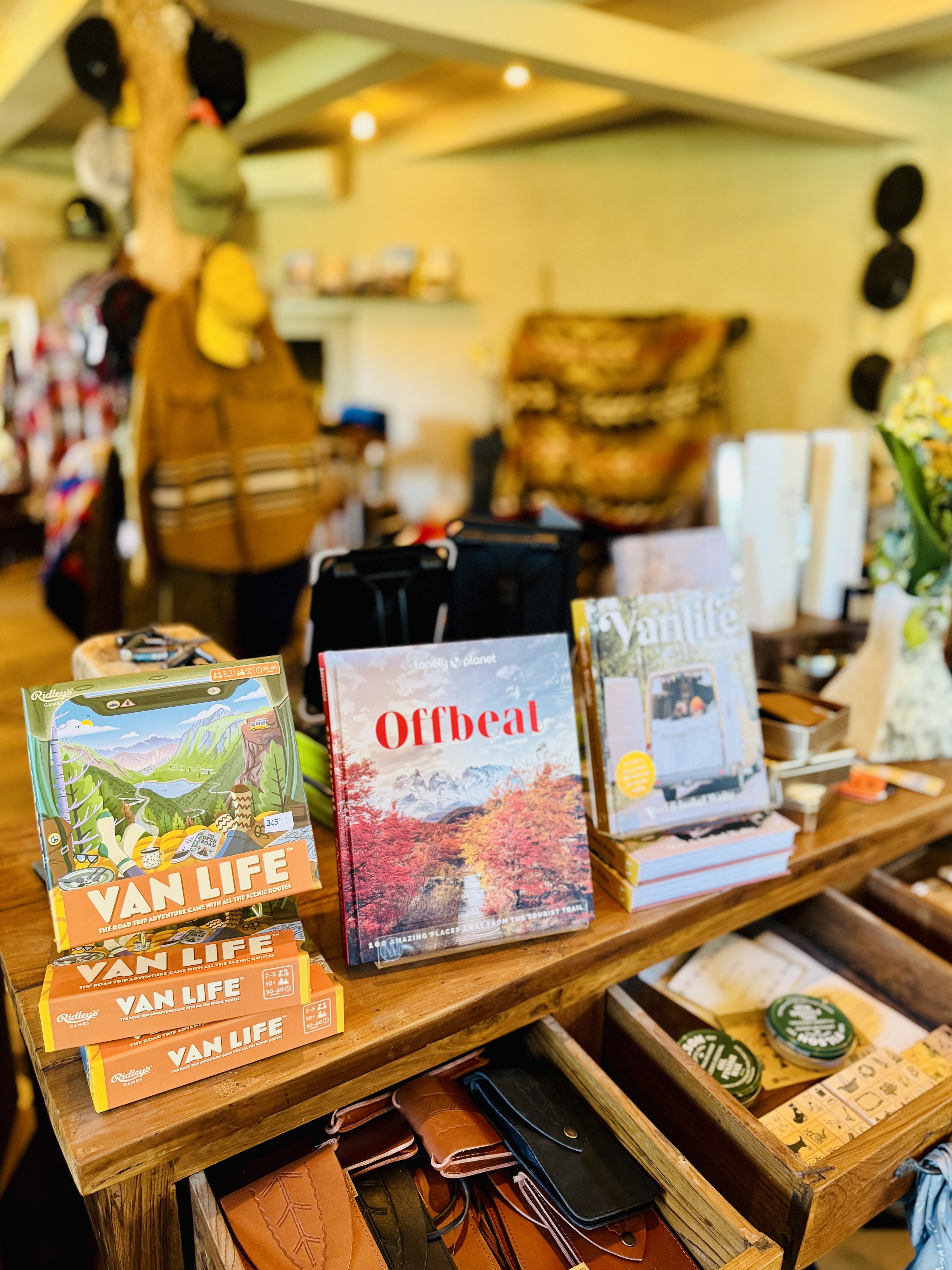 A store display featuring travel and adventure-themed books and boxes. Items include books titled "Offbeat" and "Van Life" alongside various accessories like wallets, and hats hanging in the background.