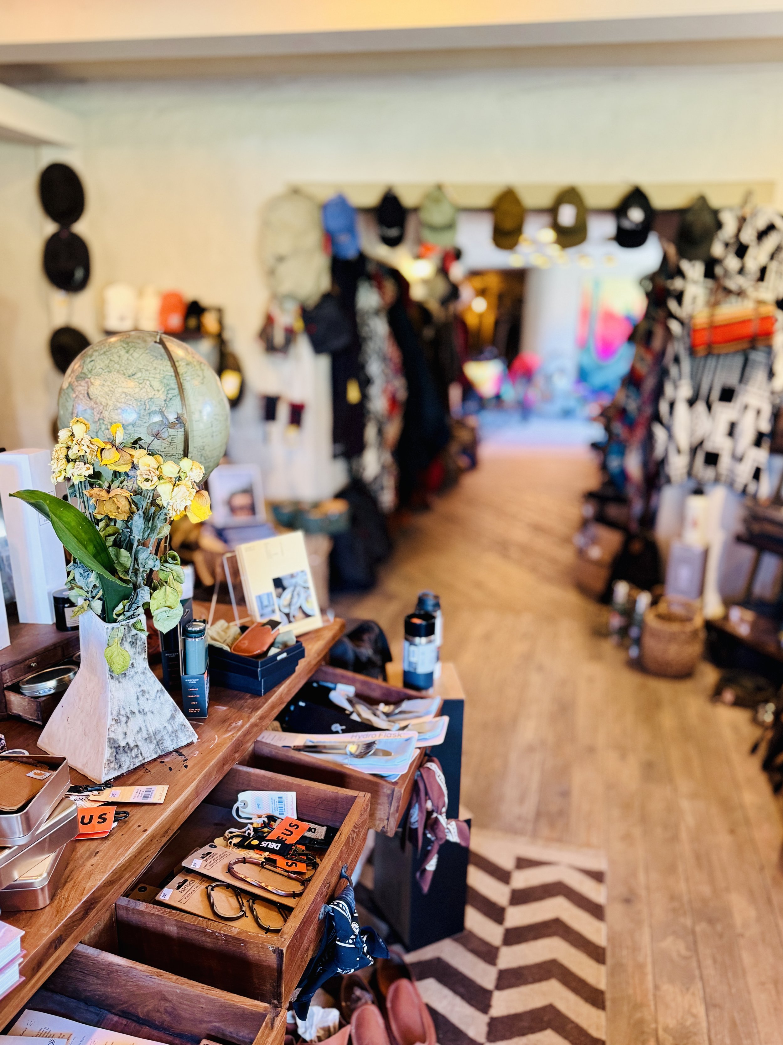 Interior of a vintage store with a wooden display table featuring a globe, dried flowers, trinkets, and accessories. Clothing and hats line the walls in the background. A chevron-patterned rug is on the wooden floor.