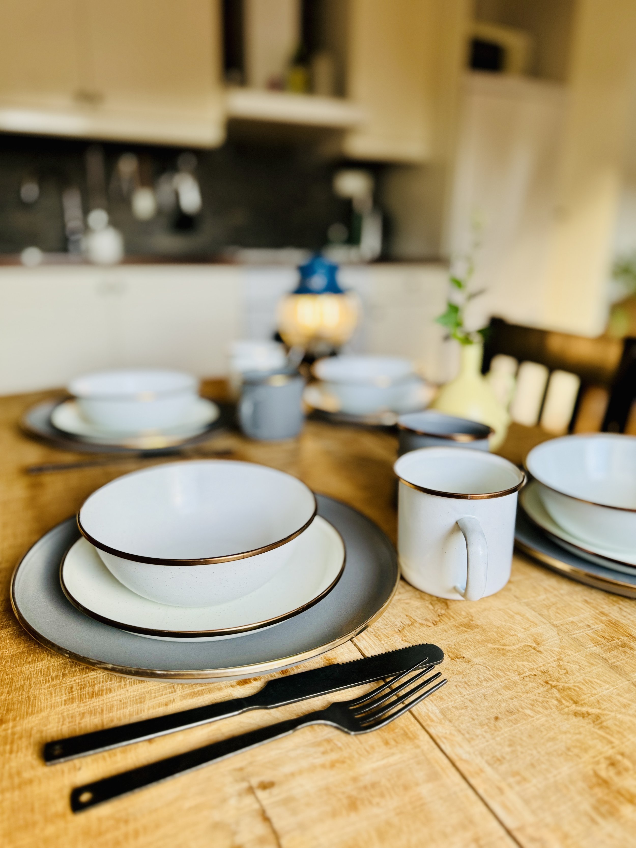 Rustic dining table set with crockery and cutlery, including plates, bowls, mugs, forks, and knives, all from Barebones in a homely kitchen environment.