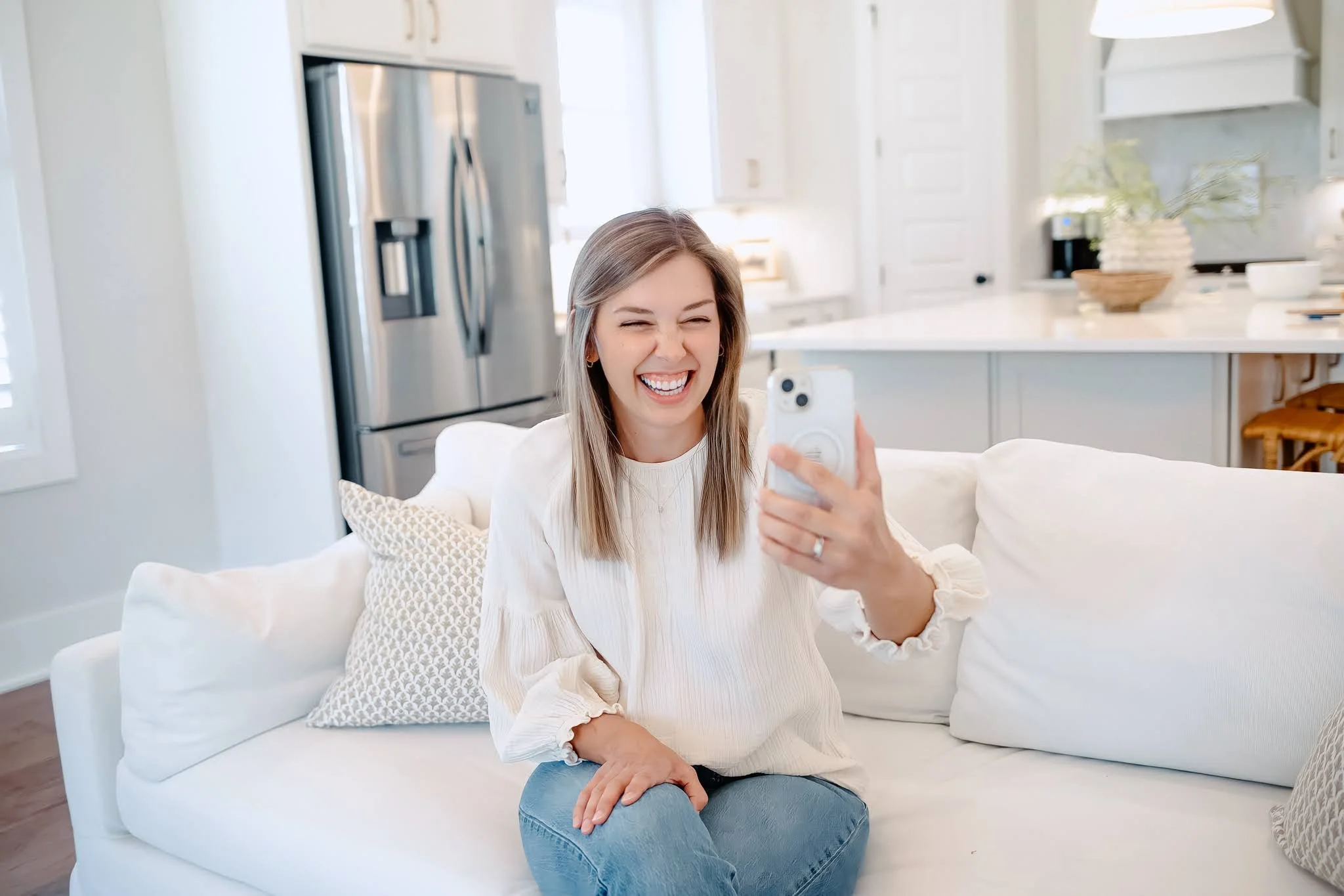 A woman sitting on a white sofa in a bright, modern kitchen, holding a phone and smiling broadly.