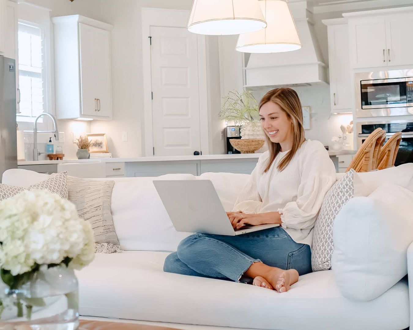 A woman sitting on a white couch using a silver laptop in a bright, modern kitchen and living room area.