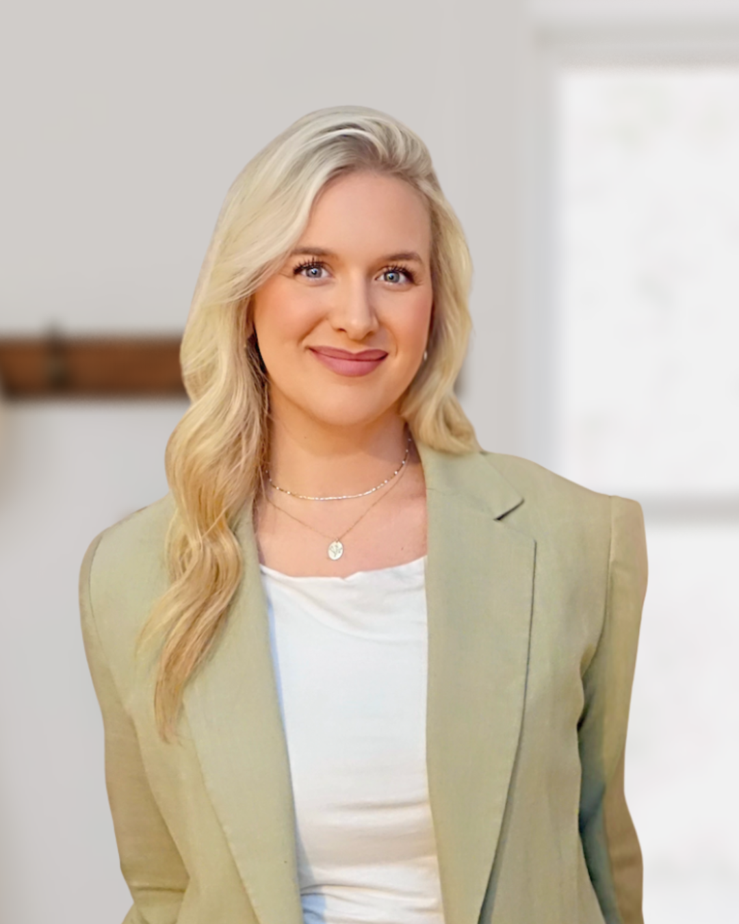 A woman with long blonde hair, wearing a light green blazer and white top, smiling indoors with a blurred background.