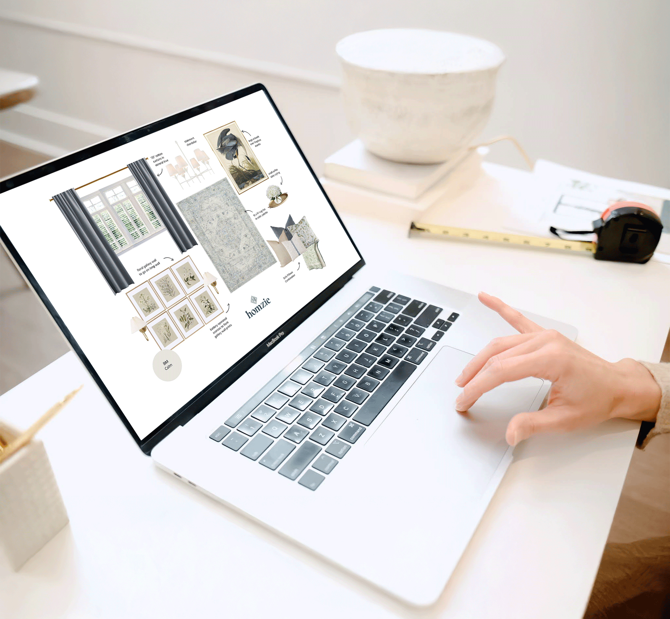 Person using a MacBook laptop with an interior design mood board on the screen, featuring curtains, wall art, and decorative pillows, on a white desk with a bowl, measuring tape, and papers nearby.