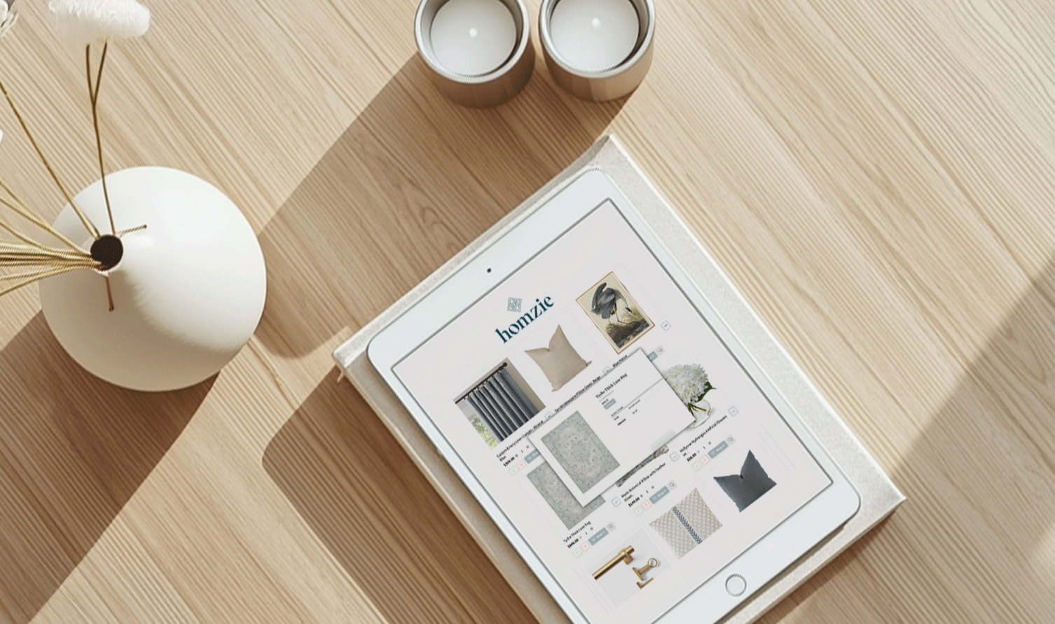 iPad displaying an interior design website on a wooden desk with a white vase holding dried plant stems and two candles.