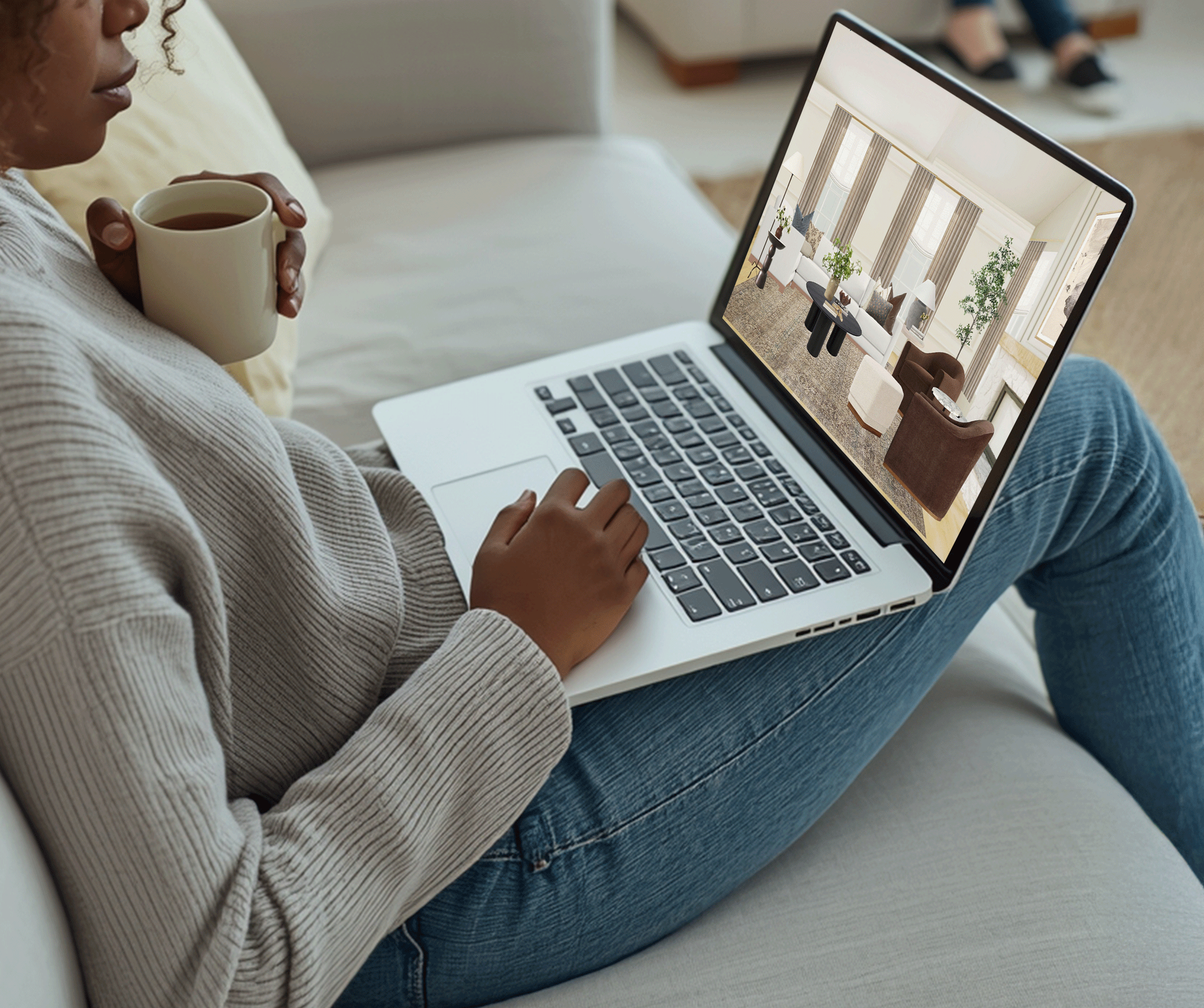 Person sitting on a sofa with a laptop on their lap displaying an interior living room design, holding a cup of coffee.