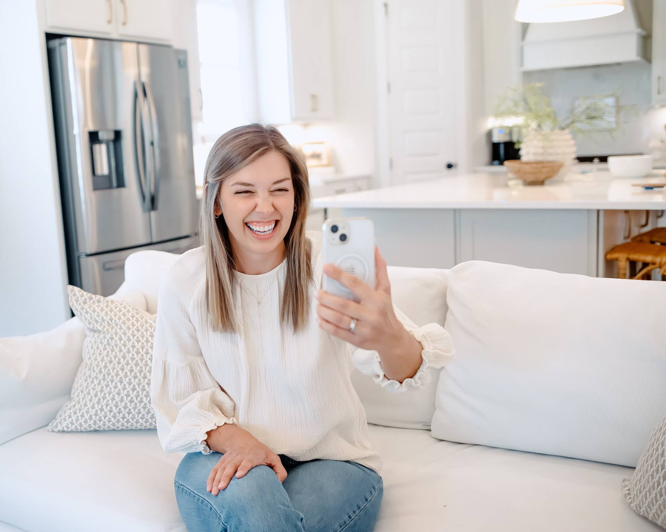 A young woman with blonde hair sitting on a white couch, laughing while taking a selfie with her smartphone in a bright, modern kitchen.