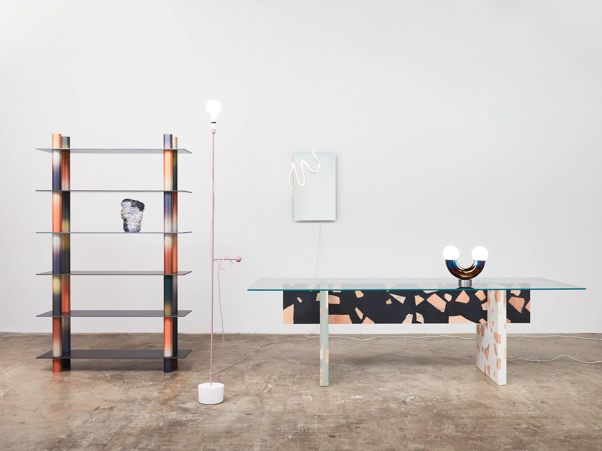 Minimalist interior with a multicolored bookshelf, a pink corded lamp, a white wall-mounted artwork, and a glass table with terrazzo-style sides, one holding a modern lamp and the other holding a decorative object.