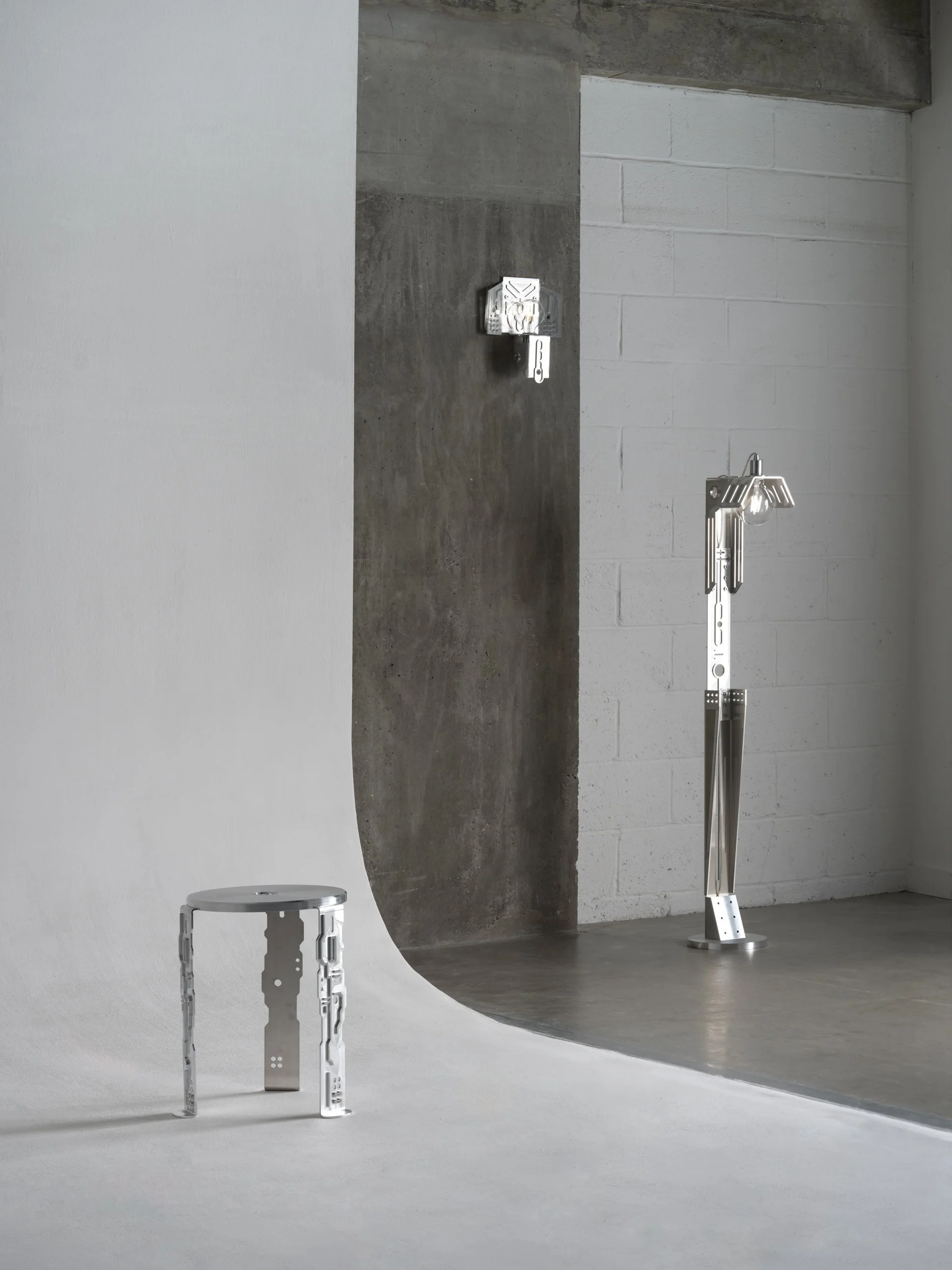 Minimalist art installation featuring metallic furniture and lighting of abstract, geometric shapes resembling mechanical parts, set against a plain background with a curved white and gray surface.