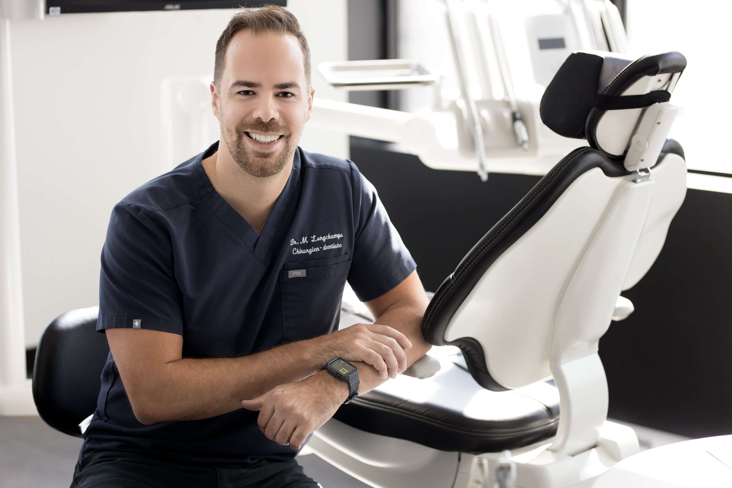 Un homme souriant en uniforme de dentiste, assis à côté d'une chaise dentaire dans un cabinet.