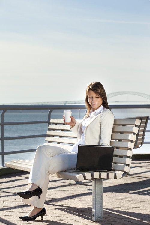 Photo - Femme assise sur un banc au bord de l'eau, tenant une tasse et utilisant un ordinateur portable