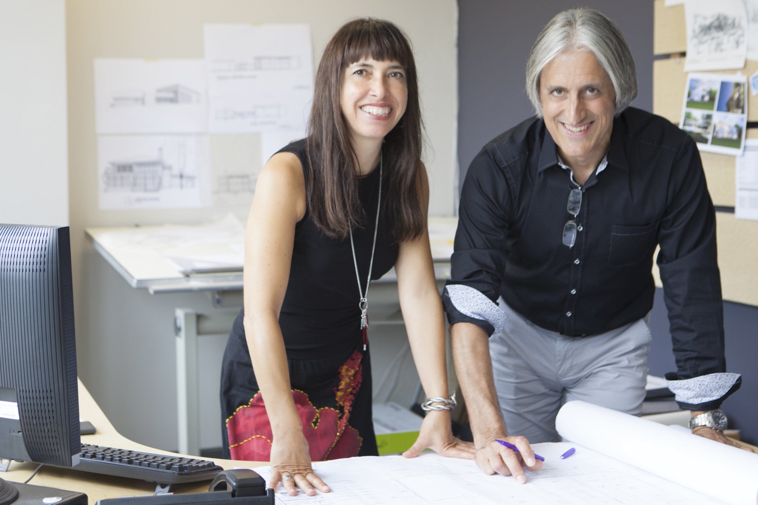 Photo de travail - Deux personnes debout à une table de travail, souriantes, dans un bureau avec des plans et des photos dans le fond.