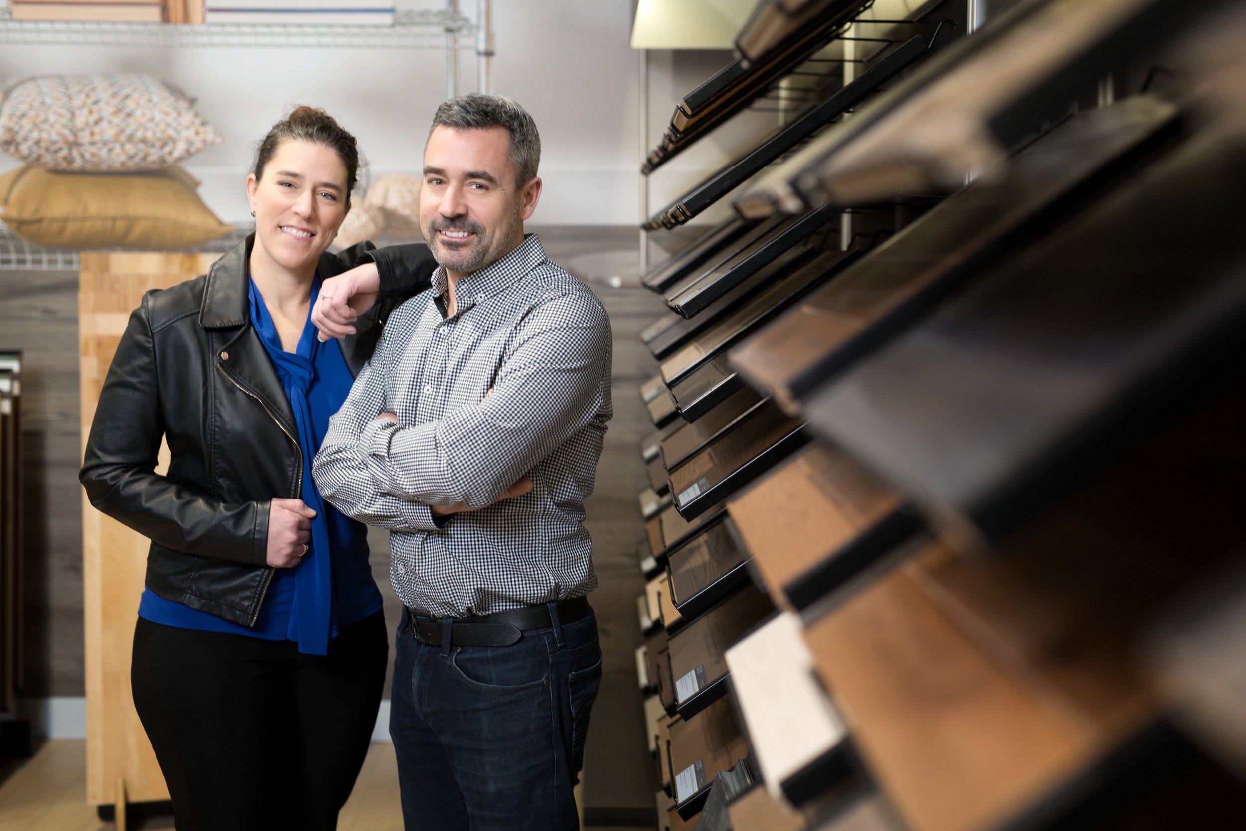 Un homme et une femme souriants posent dans un magasin de décoration intérieure, avec des échantillons de revêtements de sol en bois accrochés sur le mur à côté d'eux.