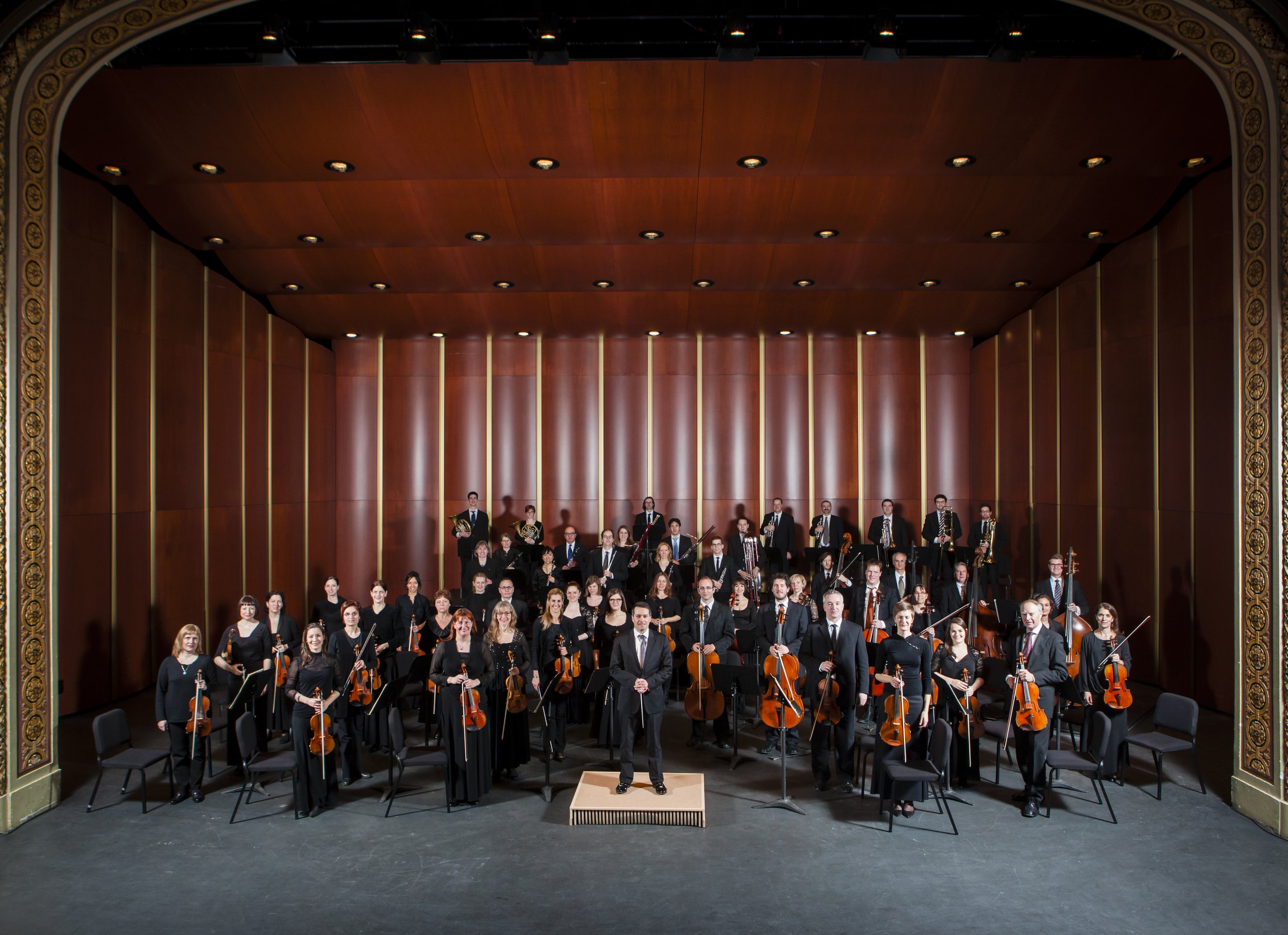 Orchestre symphonique avec musiciens et chef d'orchestre sur scène dans une salle de concert en bois.