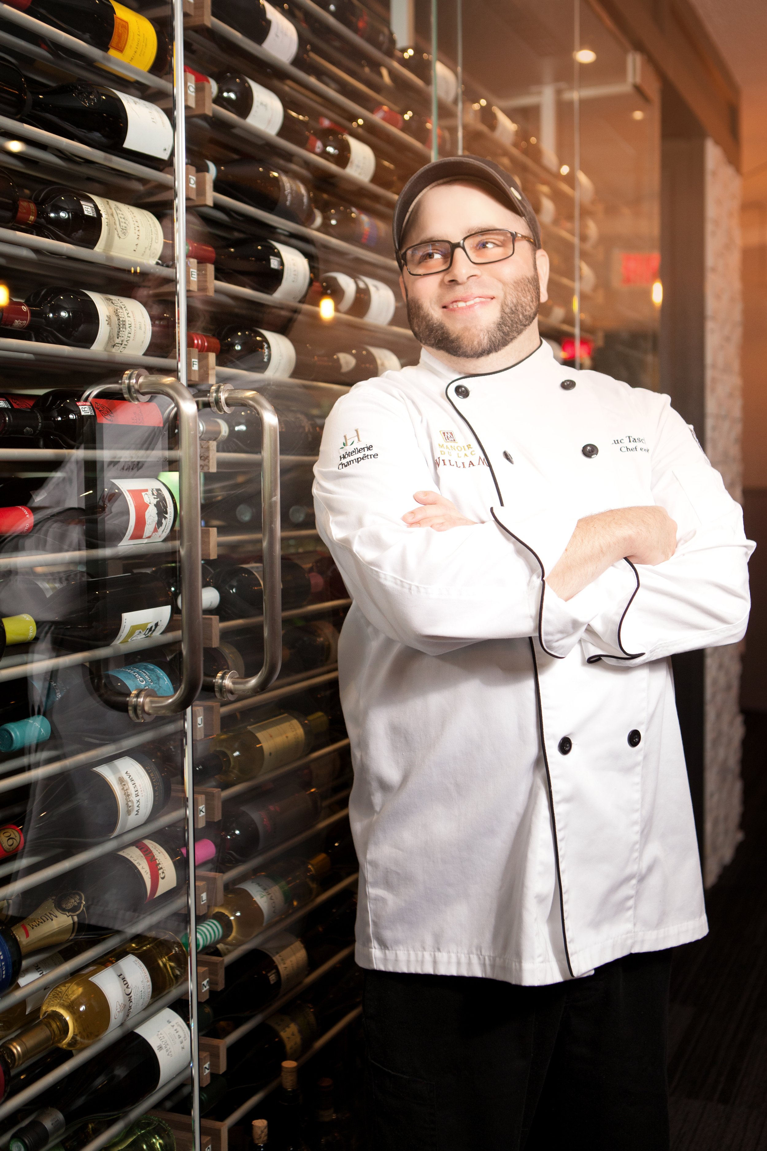 Chef professionnel posant dans une cave à vins avec des bouteilles de vin sur des étagères en verre.