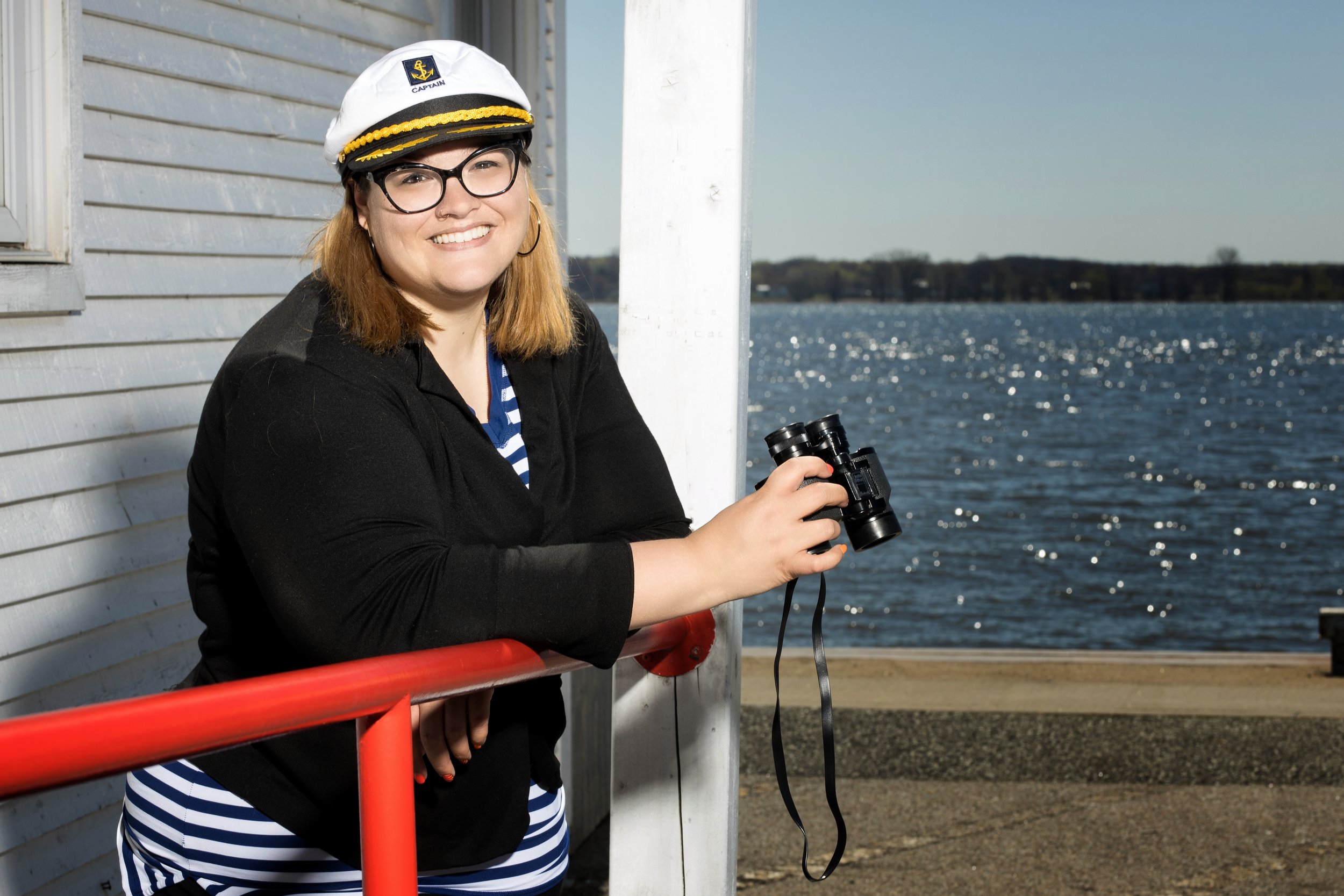 Jeune femme souriante portant un chapeau de capitaine, des lunettes, une veste noire et une robe à rayures, tenant une paire de jumelles près d'un plan d'eau, avec un bâtiment en bois à côté et un ciel clair.
