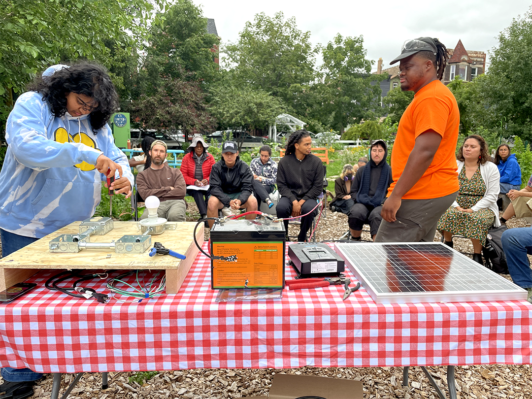 A woman preparing a science demonstration at an outdoor event with an audience seated around her, and solar panels on the table.