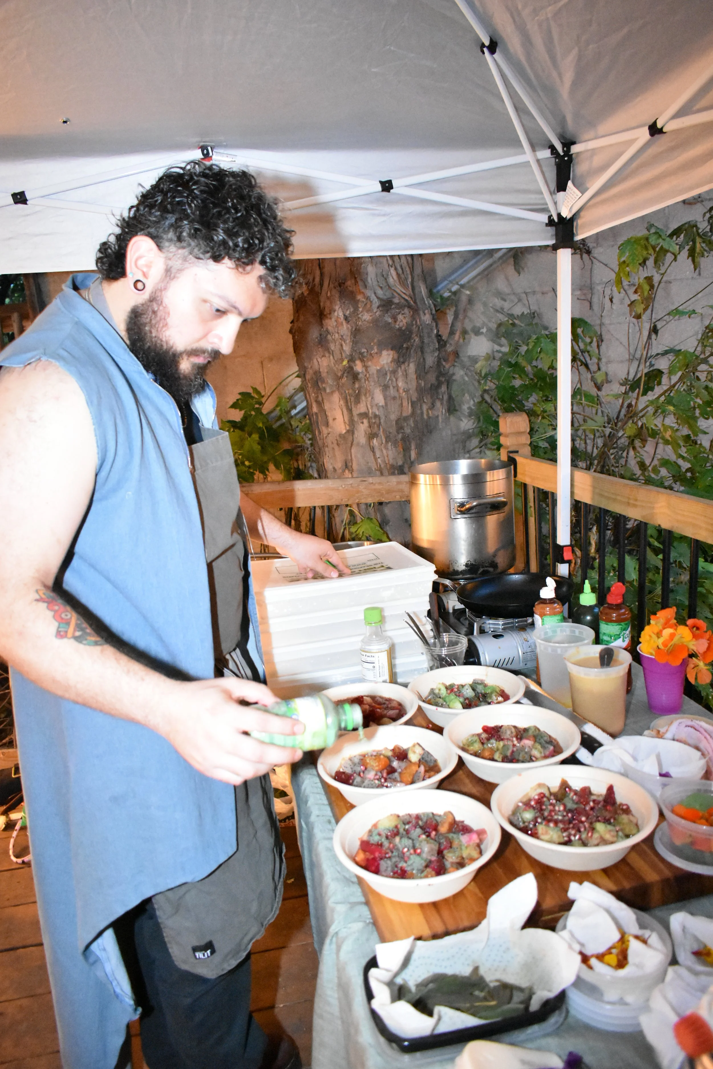 A person with curly hair and a beard, wearing a sleeveless top and apron, is preparing food at a table with bowls of salad and toppings. The setting is outdoors under a canopy, with a large tree and foliage in the background and various condiments and utensils on the table.
