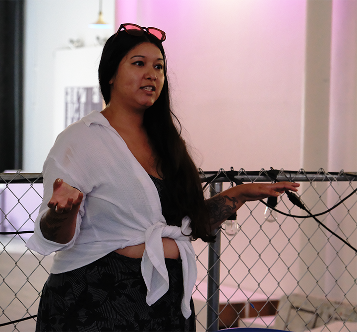 Woman speaking or presenting in front of a chain-link fence, wearing a white tie-front shirt and black patterned pants, with glasses on her head.