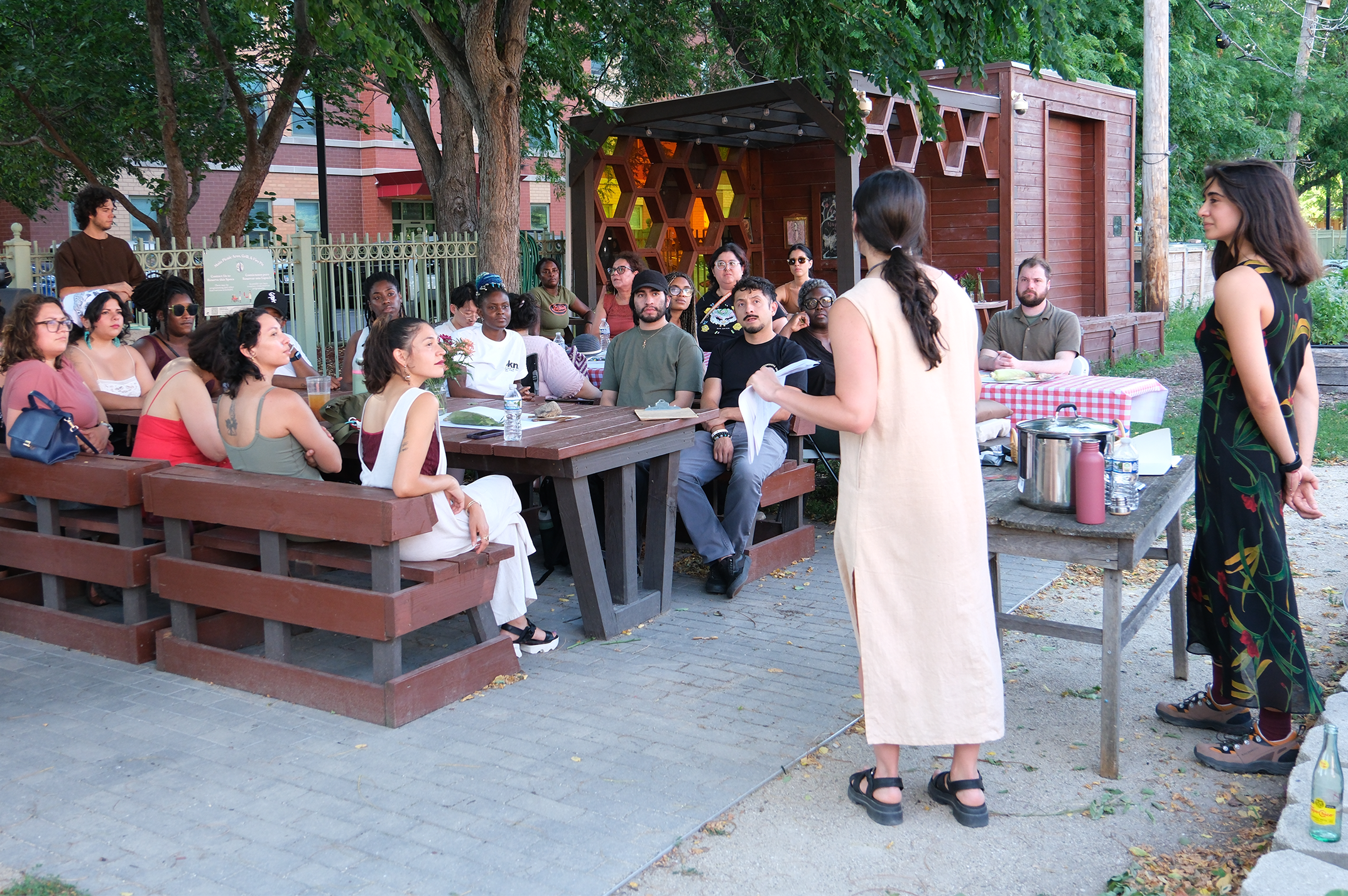 A woman with dark hair and light skin stands in front of a group of diverse people seated at picnic tables outdoors. She appears to be speaking or presenting to the audience. The setting is a park or backyard with trees and a wooden building in the background.