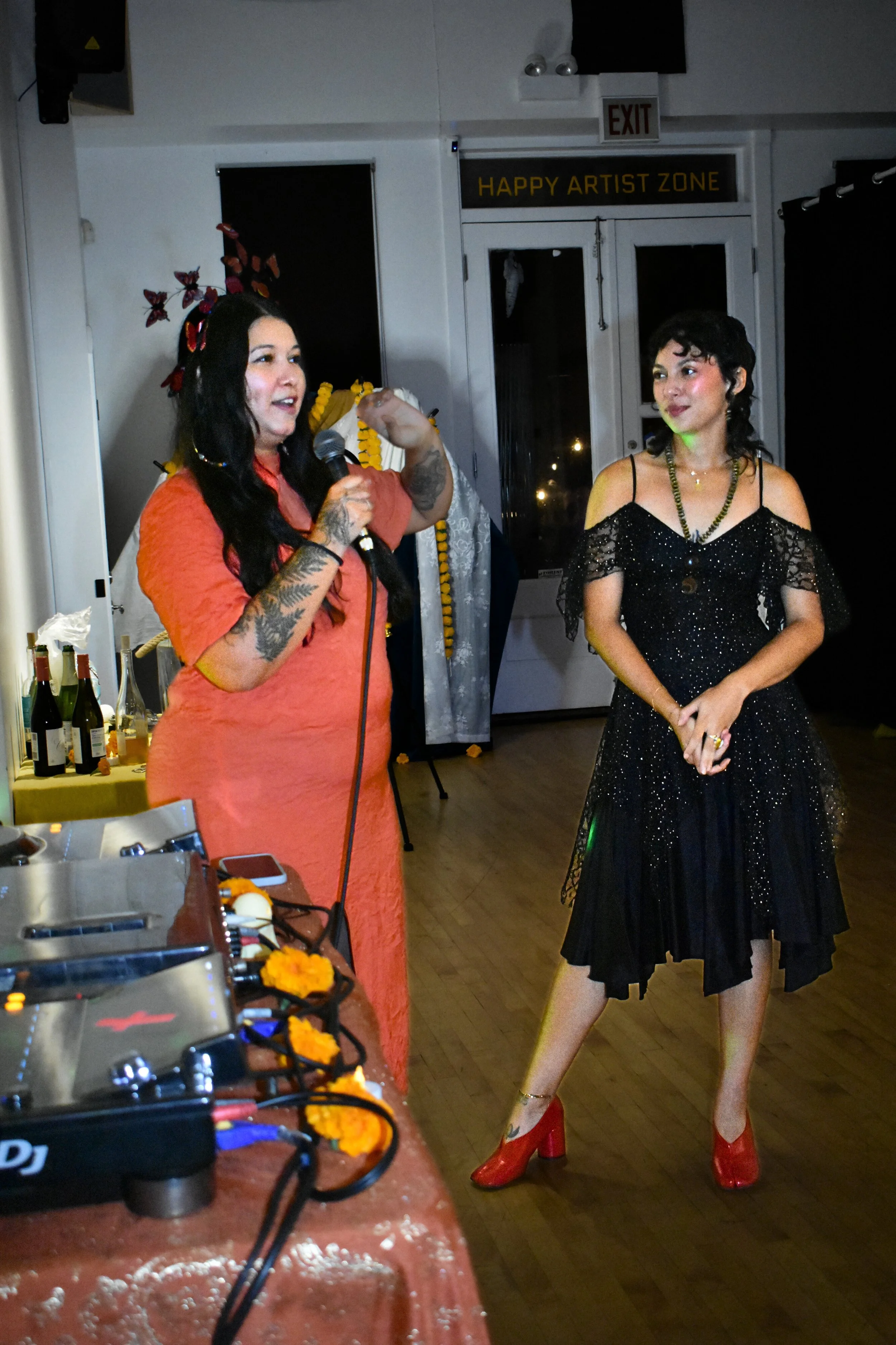 Two women standing at a social event. The woman on the left has long black hair, tattoos on her arm, a headband with red and purple butterflies, and is holding a microphone. The woman on the right has short curly black hair, wears a black sparkly off-the-shoulder dress, red high heels, and a necklace. There are bottles of wine on a table in the foreground, and a door with a sign that says 'HAPPY ARTIST ZONE' in the background.