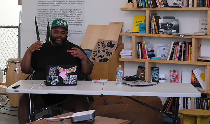 A man with a beard wearing a green cap, black T-shirt, and shorts sitting at a white folding table with a laptop decorated with colorful stickers, a water bottle, and a laptop charger. Behind him are wooden shelves filled with books and magazines, and a wall with text and a guitar. There is a drum on the left side.