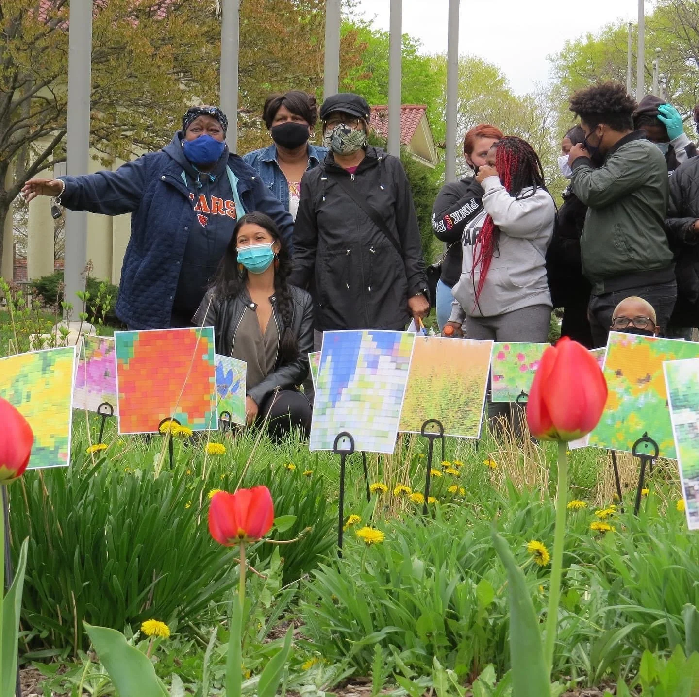 Group of diverse people gathered outdoors in a garden, wearing masks, with colorful pixelated artwork displayed among blooming tulips and yellow flowers.