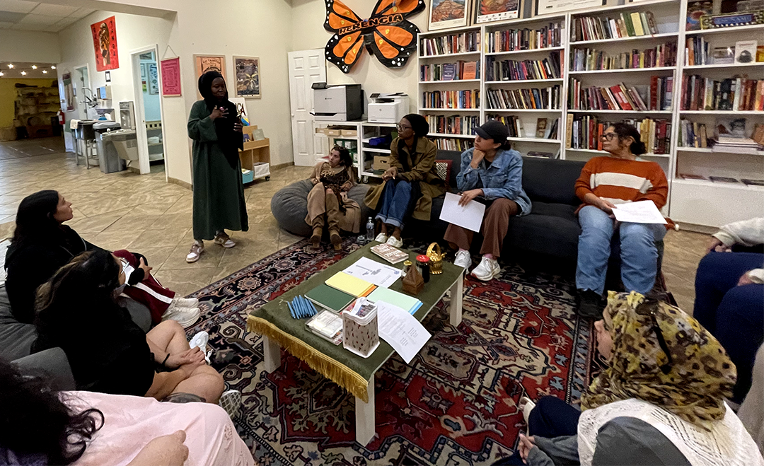 A group of people sitting and standing in a cozy room with bookshelves, listening to a woman speaking. There are papers and books on a central table, and a large butterfly decoration on the wall.
