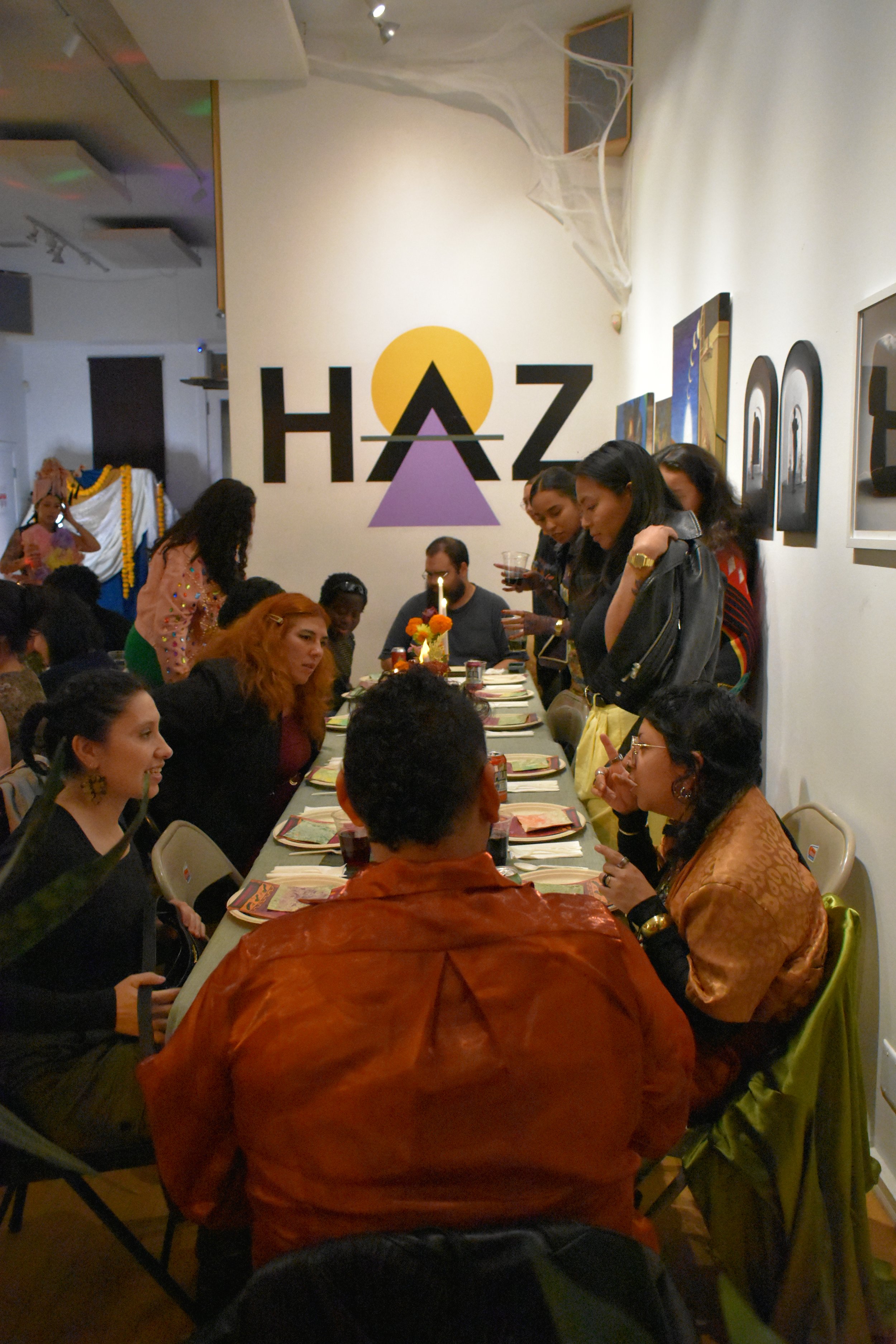 People gather around a long table with plates and drinks in an art gallery or exhibition space, decorated for Halloween with spider webs and a large wall sign reading 'HAZ' with a purple triangle and a yellow circle.