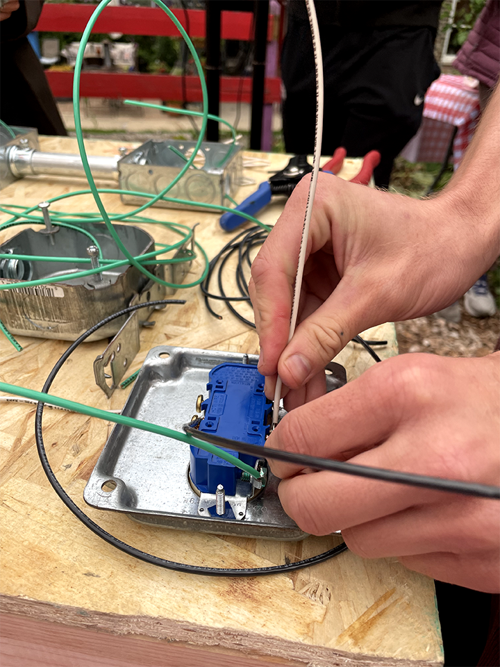 Close-up of hands wiring an electrical socket into a metal electrical box on a wooden workbench, with various wires and electrical components scattered around.