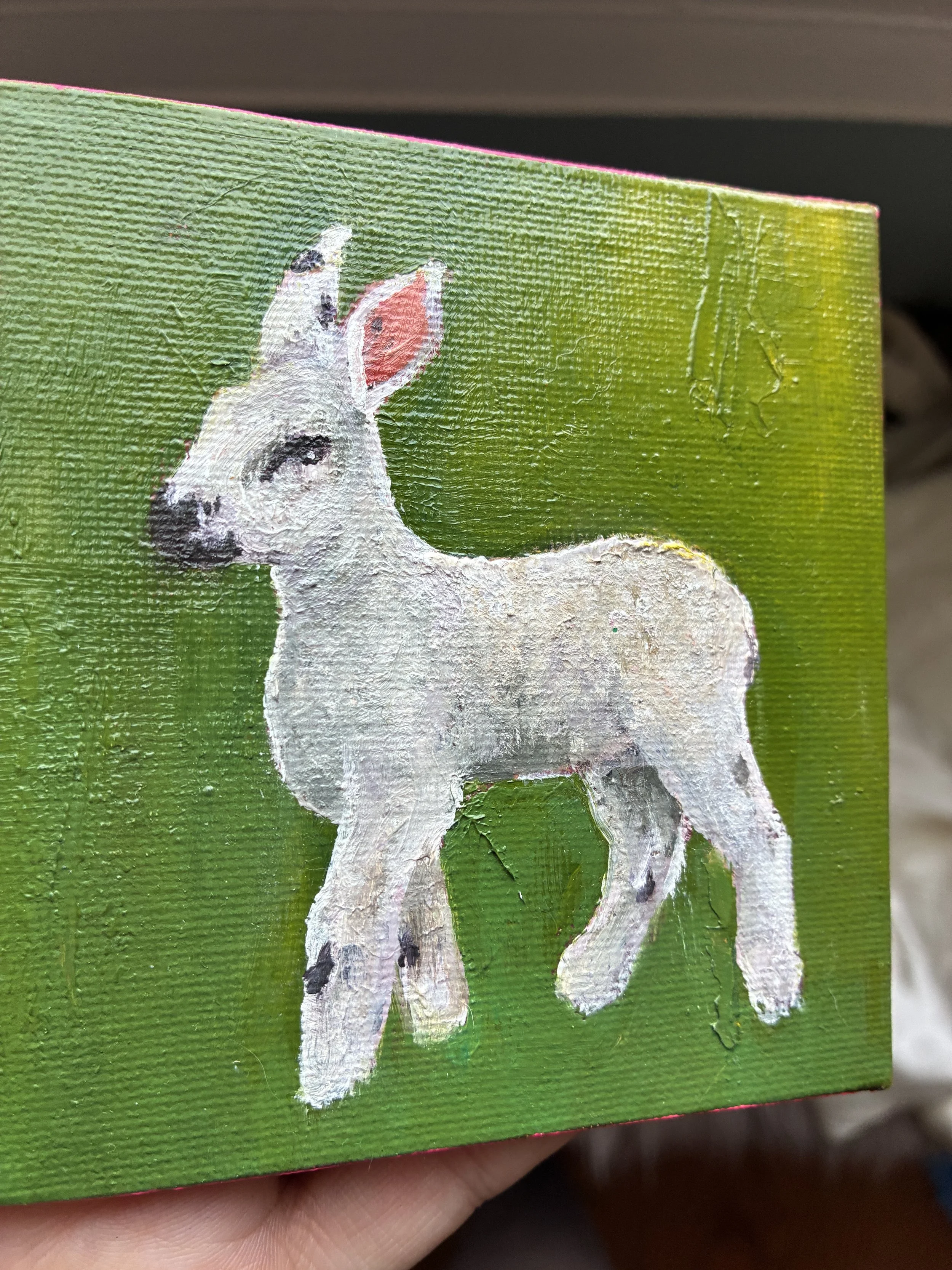 A painting of a white lamb with black spots on a green and yellow background. It is held at an angle, showing the shine on the paint.