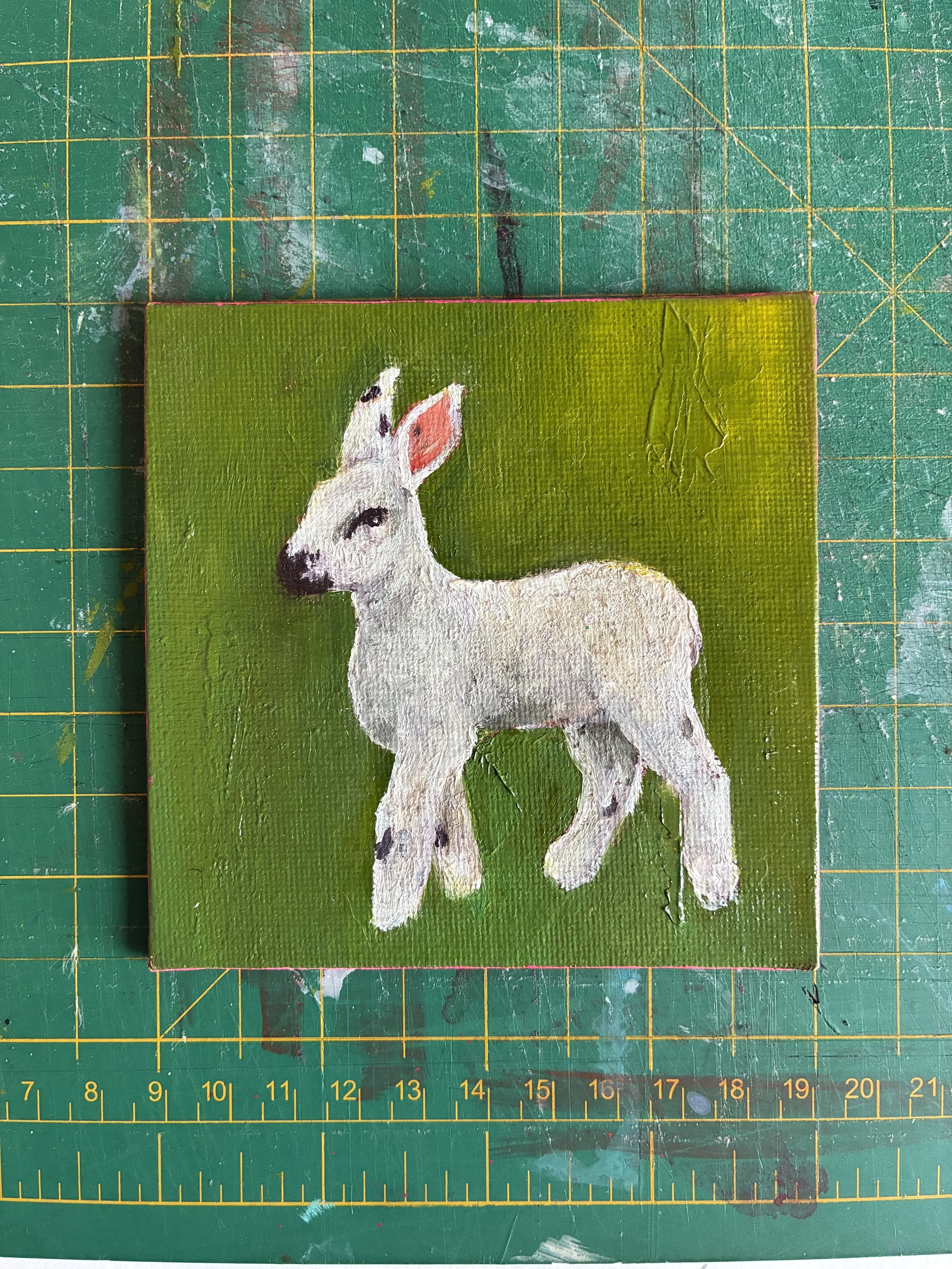 A painting of a white lamb with black spots on a green and yellow background. The painting sits on a well-used cutting mat.