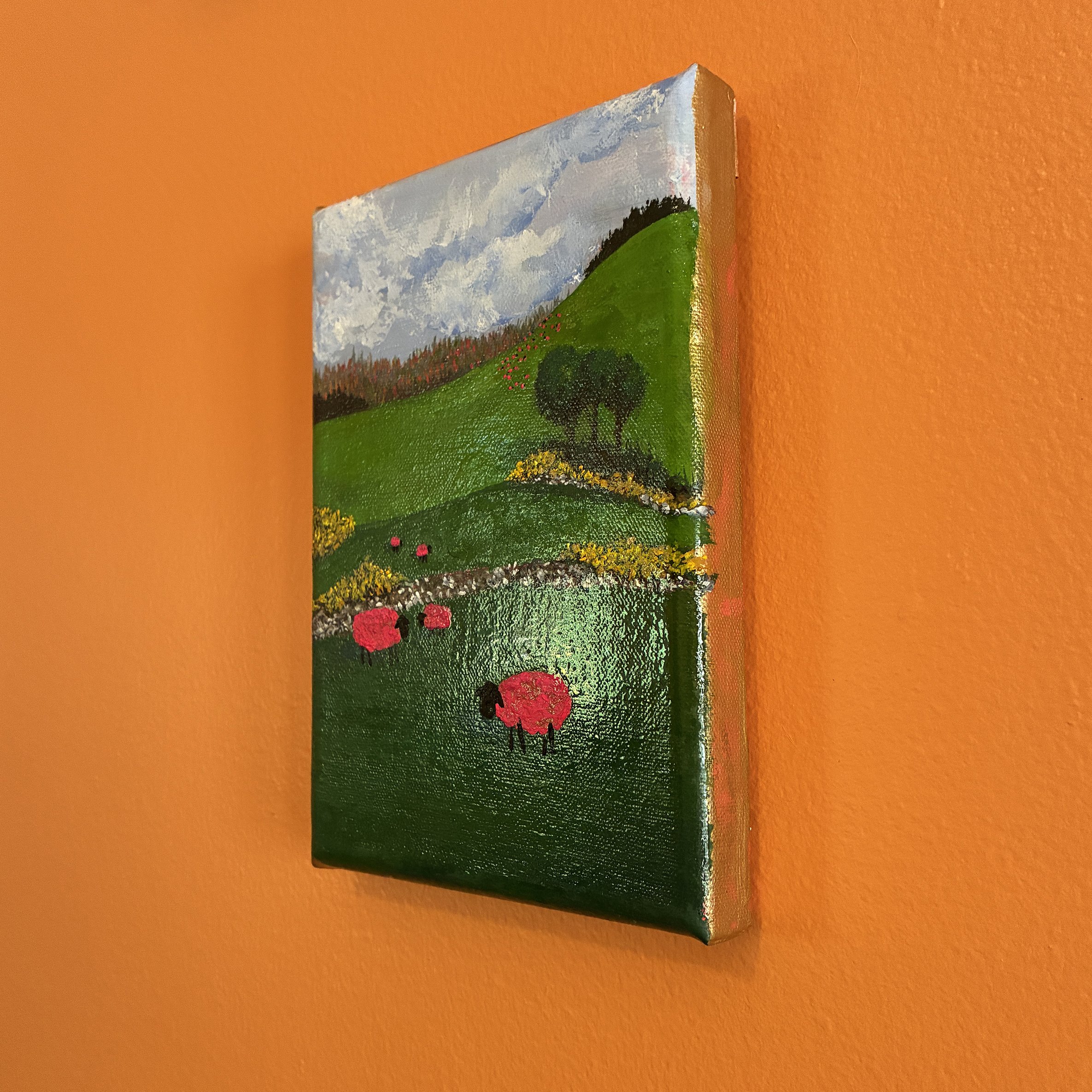 Colorful painting of pink sheep standing in green fields hanging on an orange wall. The photo shows the pink and gold edges of the painting.