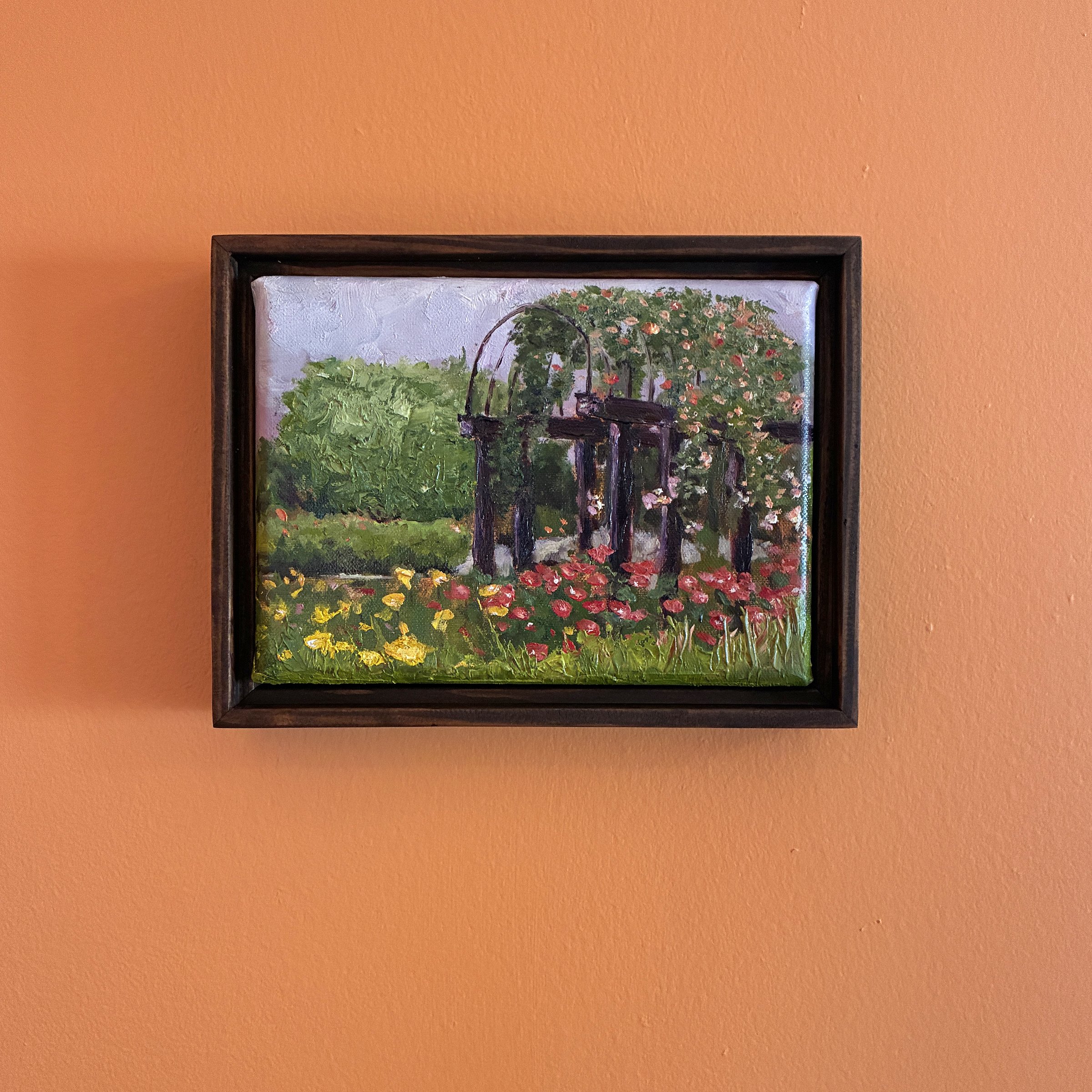 Oil painting of a pergola in a garden full of blooms, framed and hanging on an orange wall.