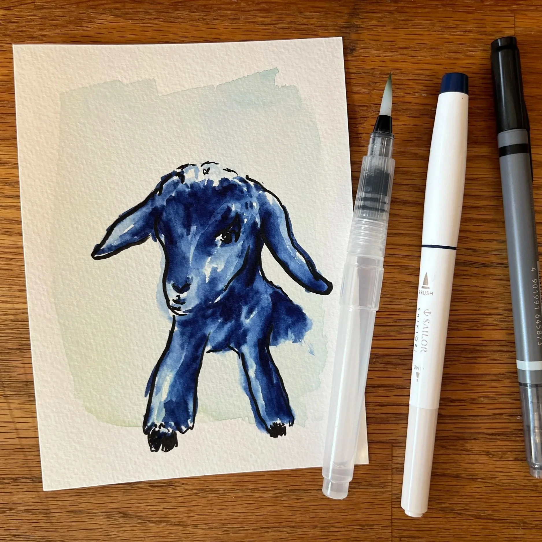 A drawing of a goat in blue ink. A water pen and markets lay next to it.