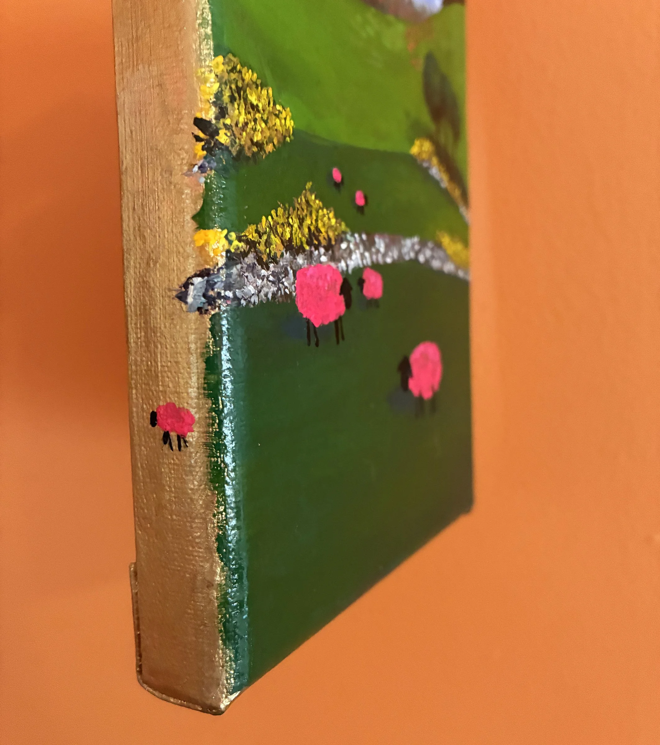 Colorful painting of pink sheep standing in green fields hanging on an orange wall. The photo is at an angle, showing one pink sheep on the gold edge of the canvas.