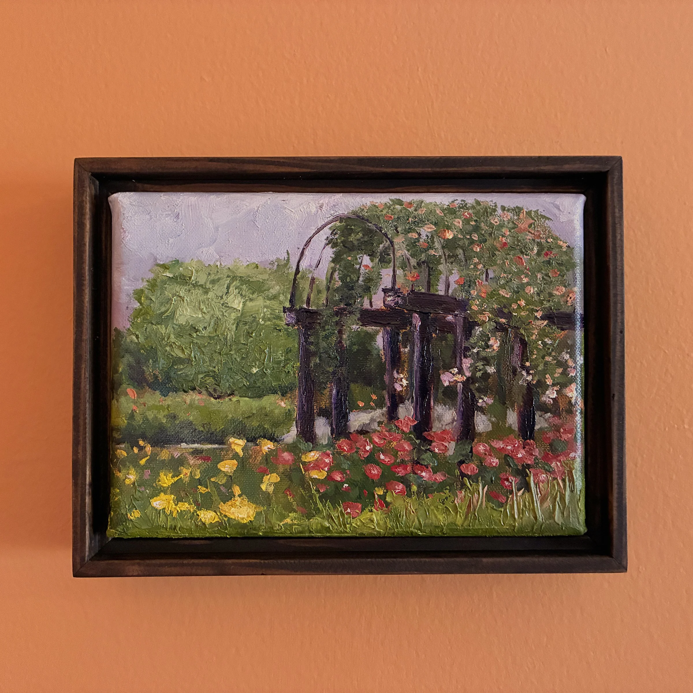 Oil painting of a pergola in a garden full of blooms, framed and hanging on an orange wall.
