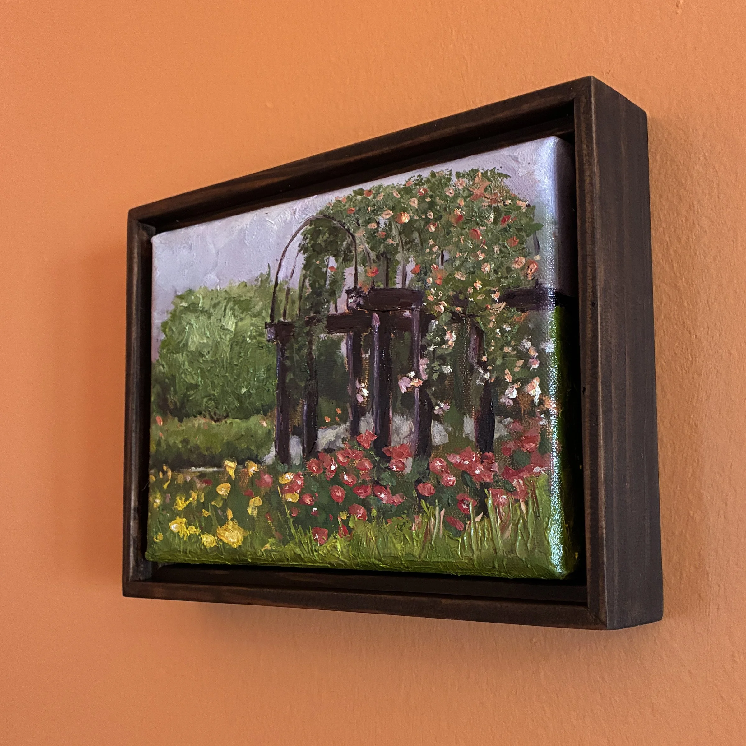 Oil painting of a pergola in a garden full of blooms, framed and hanging on an orange wall.
